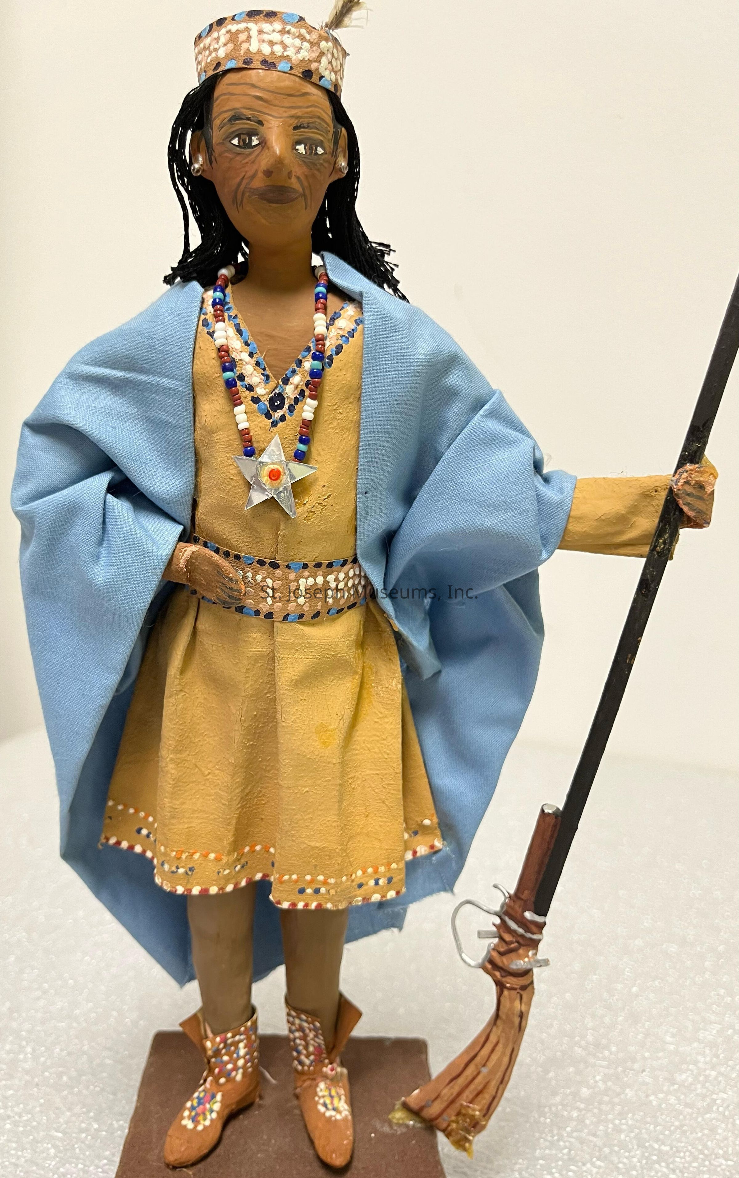 The image depicts a handcrafted doll with a detailed, culturally-inspired design. The doll wears an elaborate costume that includes a beige dress with multicolored beadwork on the edges and accessories, such as a necklace, that enhance its intricate appearance. The doll is adorned with a light blue cape draped gracefully over its shoulders. The headpiece matches the dress with similar bead patterns and a feather, further adding to its distinctive look. The figure is holding a staff in one hand, indicating a possible ceremonial or symbolic role. The boots feature detailed designs akin to the dress, with small colorful dots that provide a cohesive look. The craftsmanship suggests a blend of traditional artistic elements, reflecting cultural heritage or storytelling themes. The expression on the doll's face is carefully painted, featuring significant detail around the eyes and mouth, giving it an expressive demeanor. The entire composition sits upon a simple base, suggesting stability and display purposes, making it not only an artistic creation but also a possible collector's item or cultural artifact.