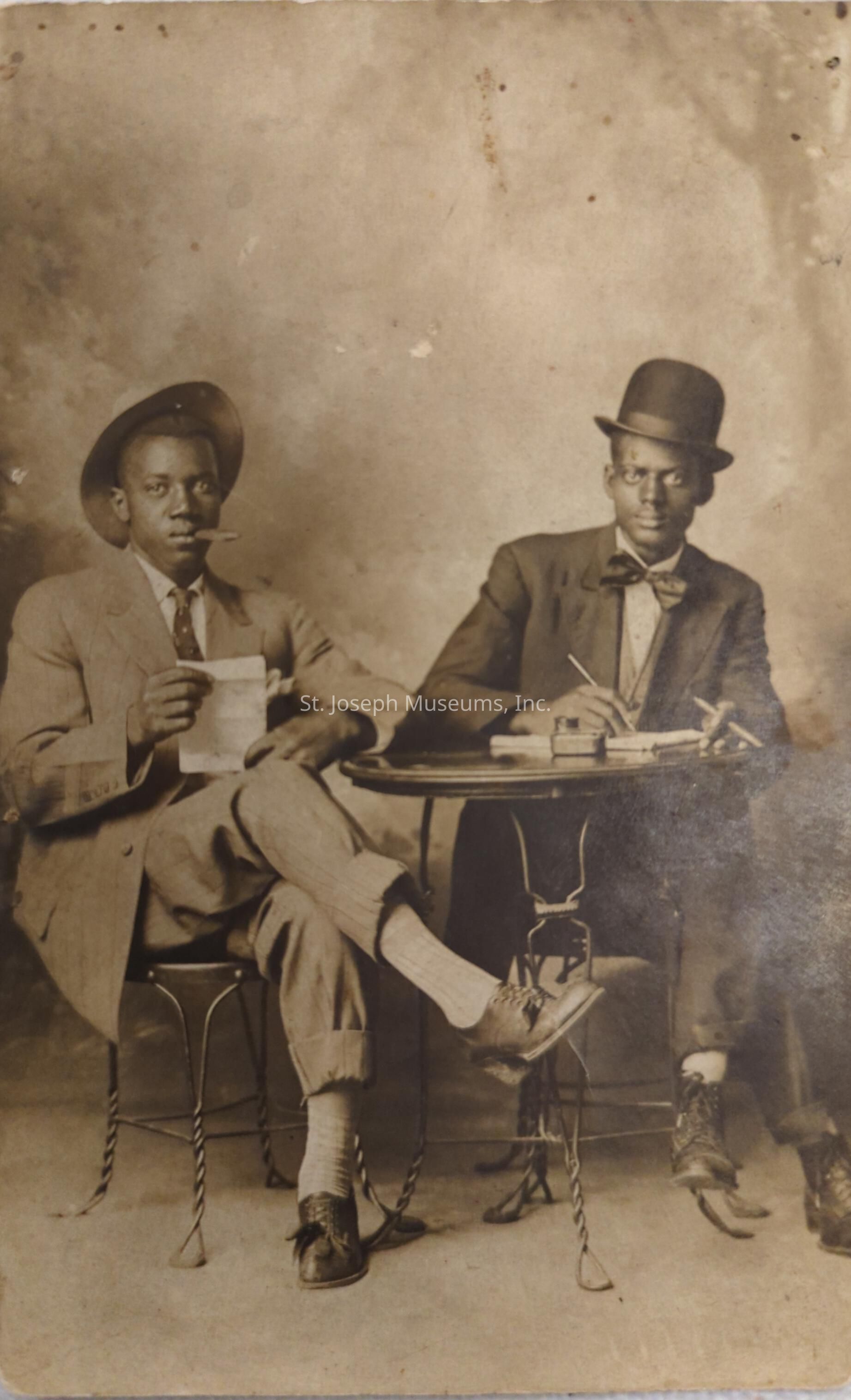 In the sepia-toned photograph, two well-dressed black men sit at a small round table with twisted metal legs, exuding confidence and sophistication. The man on the left, wearing a neatly tailored suit, a polka dot tie, and a fedora hat, casually holds a cigar in his mouth and a piece of paper in his hand while crossing his legs in a relaxed yet assertive posture. His companion, on the right, dons a bowler hat and bow tie, his serious expression focused on the task at hand as he writes with a fountain pen on paper spread across the table, accompanied by an ink pot. The backdrop, a textured wall, enhances the vintage ambiance of the scene, providing an understated and neutral canvas that highlights the subjects' dignified presence. Their attire, marked by rolled-up pant cuffs revealing white socks, adds an element of uniqueness, reflecting both the era's fashion and personal flair. This candid moment captures not only their style but also an intriguing sense of purpose.