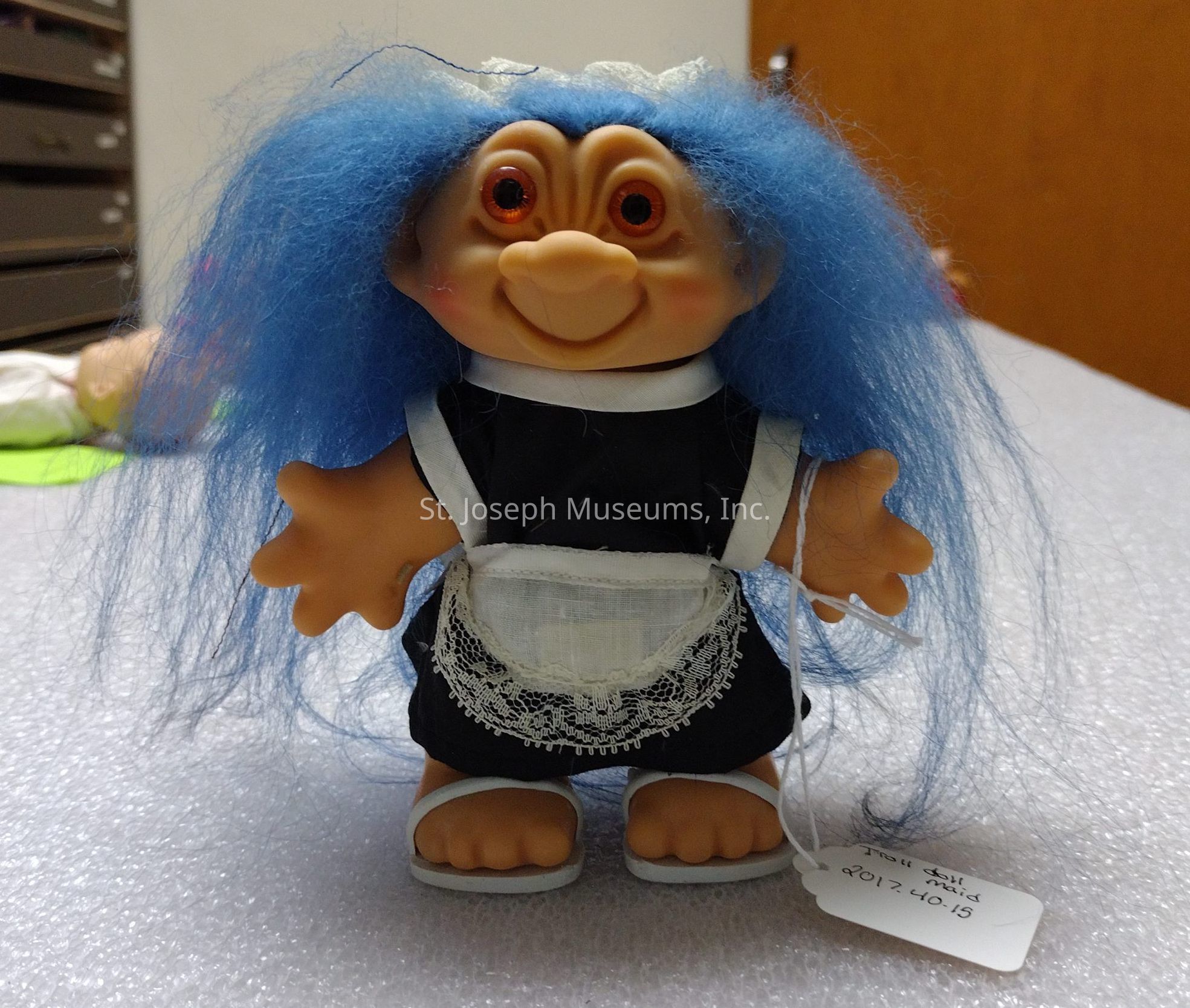 The image shows a troll doll with bright blue hair, large orange eyes, and a cheerful expression, wearing a black and white maid outfit complete with a white lace apron. The doll is standing upright on a light-colored, textured surface, and it has a tag attached to its hand that reads "Troll Maid 2012 40-15." The background includes indistinct elements, like a wooden door on the right and some drawers on the left, slightly blurred, which make the troll doll the central focus of the picture. The unique combination of the doll’s colorful hair and maid attire gives it a whimsical and playful appearance.