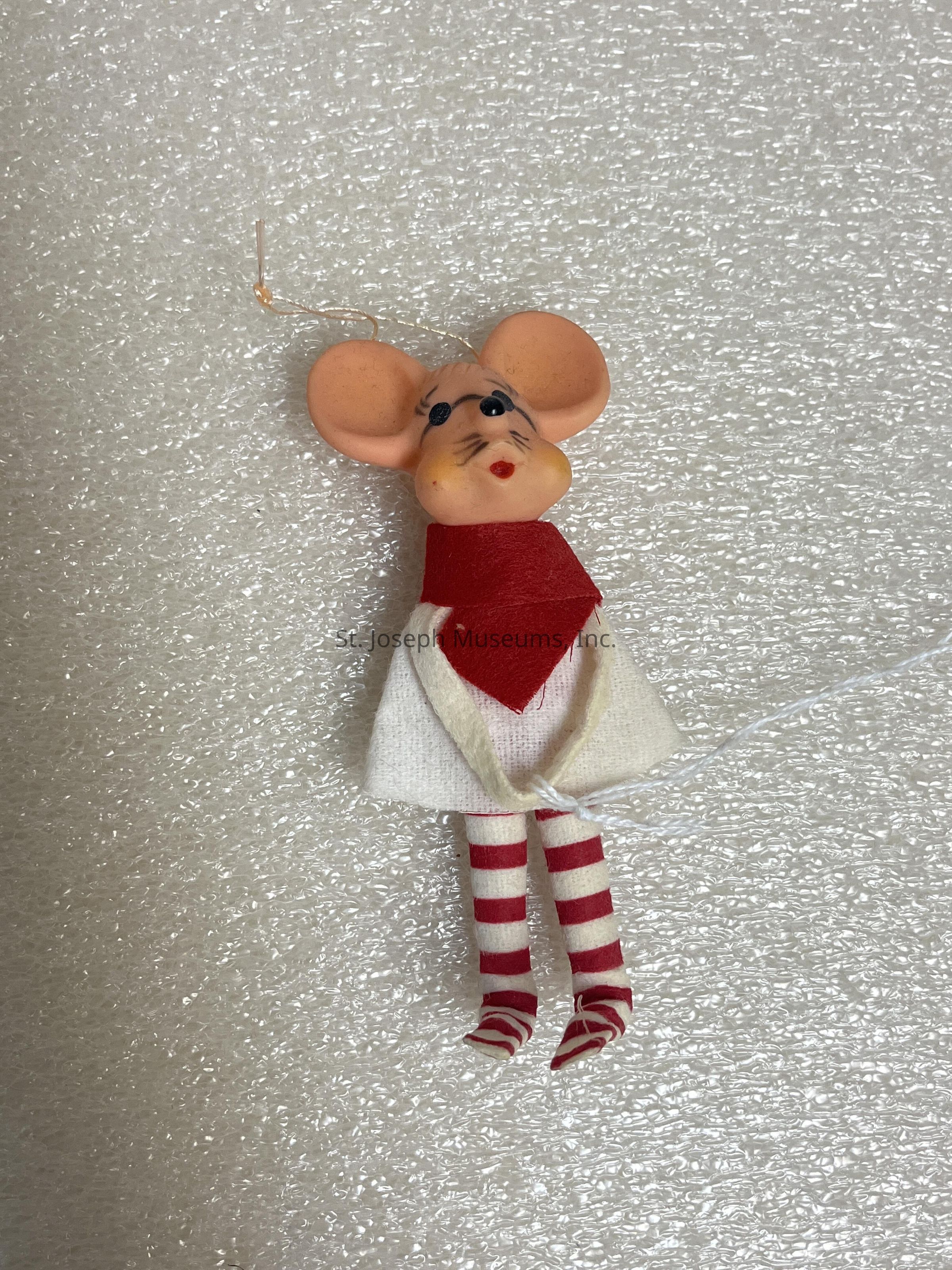 The image shows a whimsical ornament resembling a mouse with oversized ears and a painted face, featuring black eyes and whiskers. It is outfitted with a white dress accompanied by a bold red scarf tied around its neck, and its legs are adorned with red and white striped stockings. The mouse ornament hangs from a thin gold piece of wire attached to the top of its head, suggesting it could be suspended as a decoration. The entire figurine rests on a textured, white, polystyrene foam background, highlighting its festive appearance and intricate craftsmanship, making it an eye-catching and charming piece likely suitable for holiday decoration or a playful decorative theme.