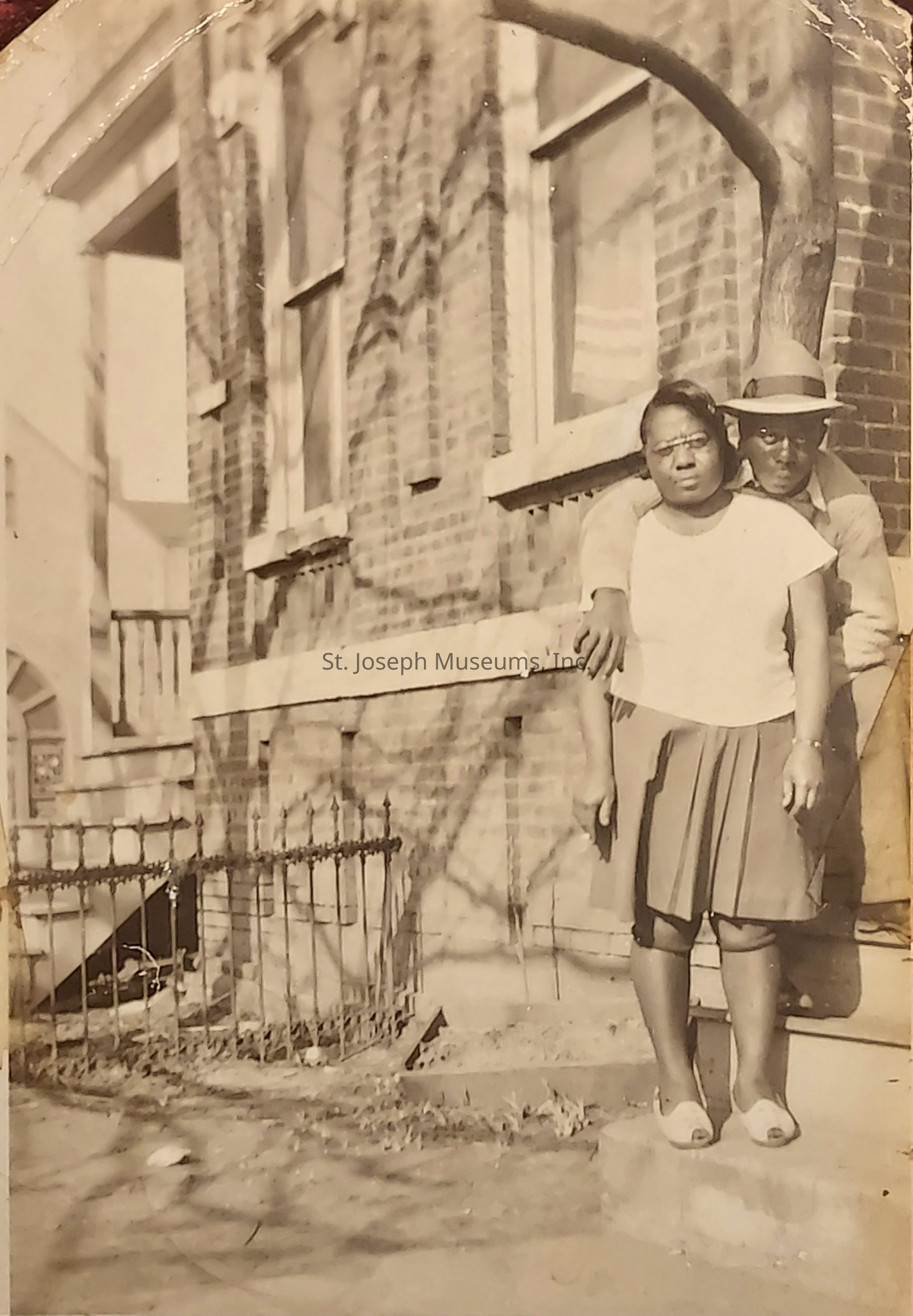 The sepia-toned image depicts a couple standing closely in front of a brick building. The man, wearing a hat and glasses, stands behind the woman with his arms draped over her shoulders. She wears a white top and pleated skirt, and appears slightly serious as she looks toward the camera. The shadows of branches play across the wall, adding texture to the scene. An old iron fence and steps leading to a doorway are visible on the left side. The overall atmosphere evokes a sense of history and intimacy, capturing a moment of connection between the two individuals against an urban backdrop.
