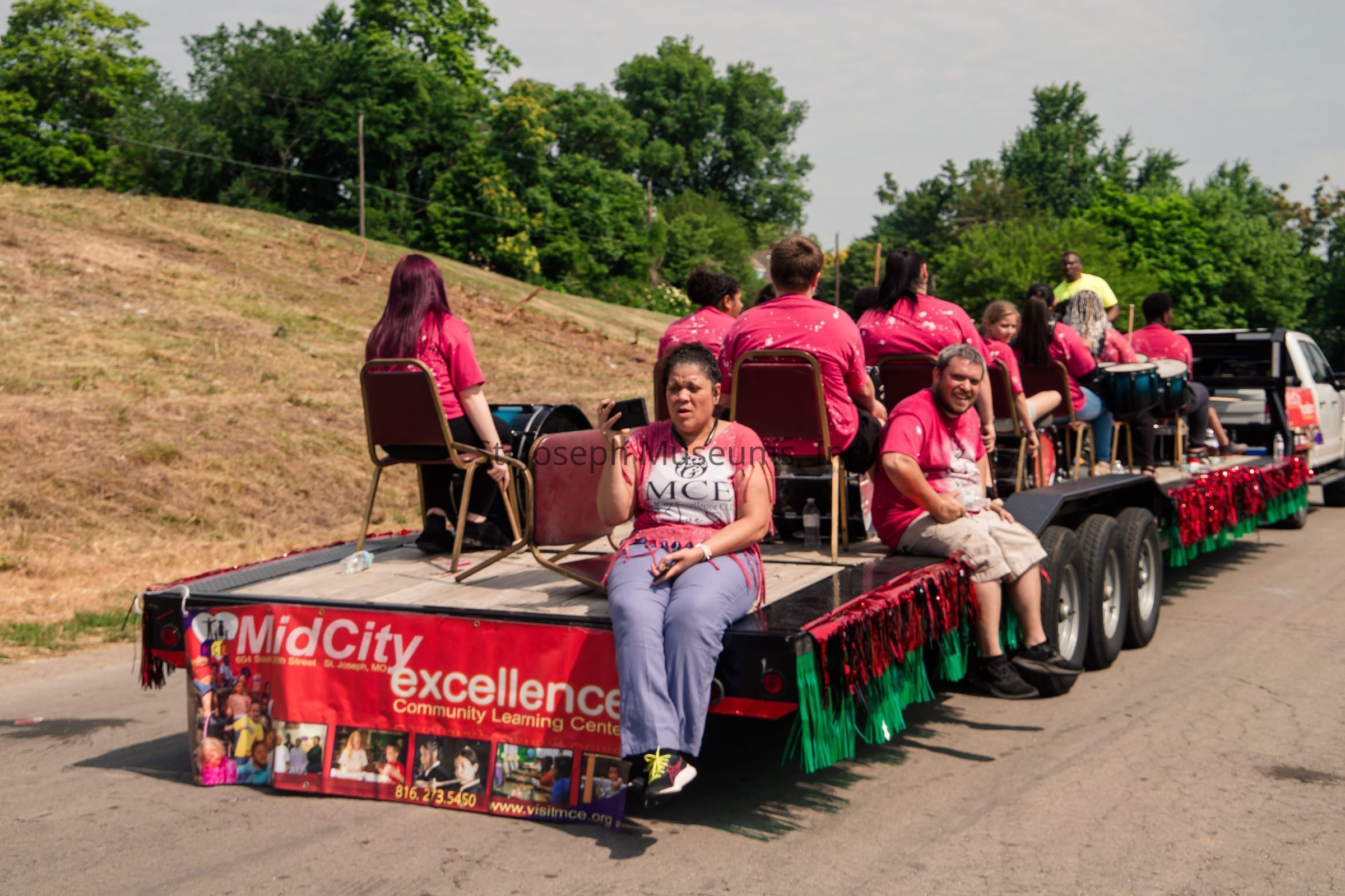 The image depicts a parade float adorned with festive red and green fringe, carrying several individuals seated in chairs. The front of the float features a large banner that reads "MidCity Excellence Community Learning Center," promoting a local organization. Most of the people are wearing bright pink shirts with similar design patterns, indicating they are part of the same group. In the foreground, a woman dressed in a shirt with "MCE" written on it is looking at her phone, while another person next to her appears to be smiling at the camera. The background shows a grassy area with lush green trees under a partly cloudy sky.