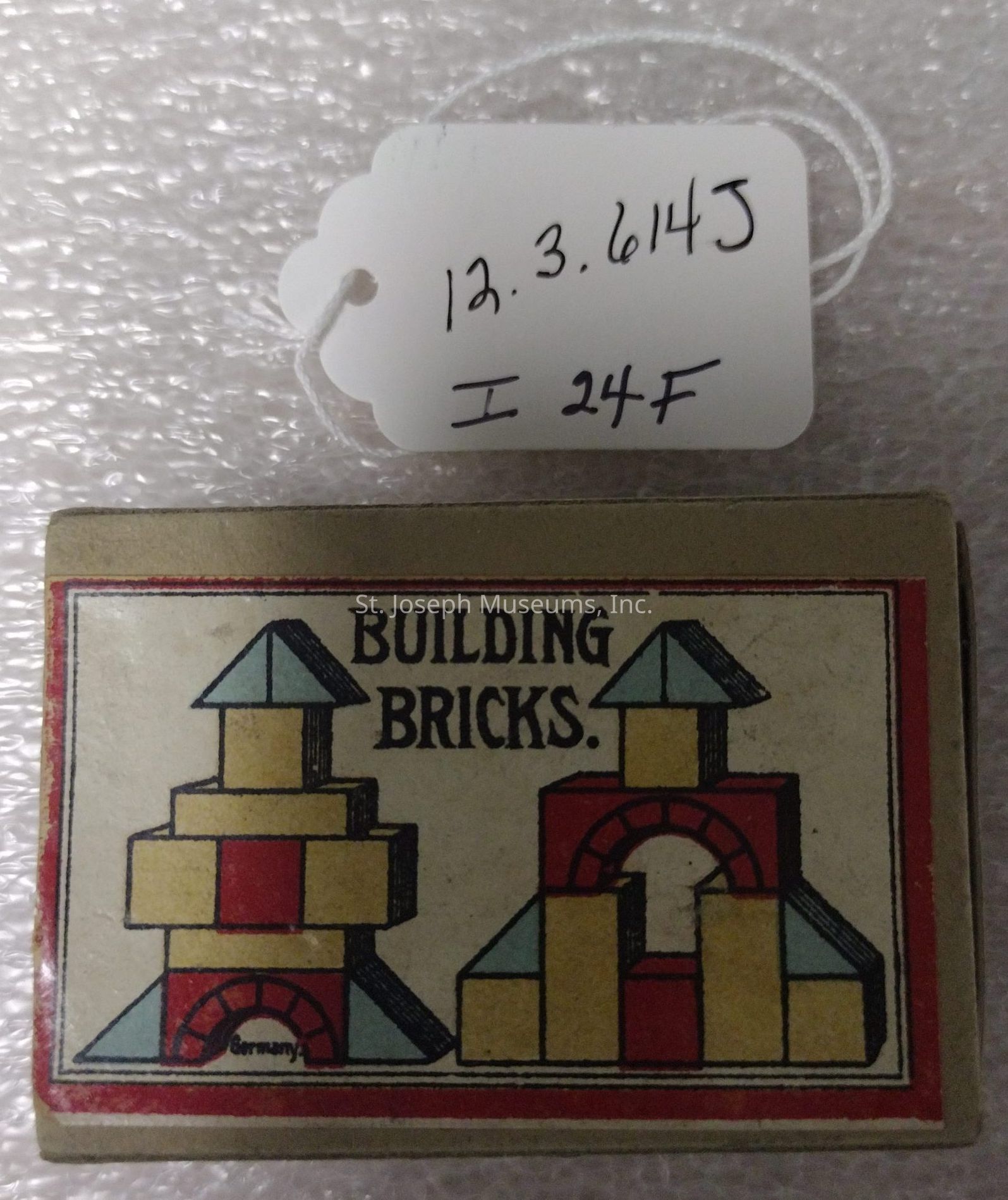 The image features a vintage packaging for "Building Bricks," likely a toy set from Germany, as indicated by the label printed on the box. The design includes colorful geometric shapes assembled into stylized constructions, primarily using red, yellow, and blue tones, with black outlines giving definition to the forms. The background is a textured surface suggesting a vintage or retro aesthetic. The box is topped with a handwritten label attached by string, displaying the numbers "12.3.614J" and "I 24F," possibly indicating catalogue numbers or inventory details. The overall appearance suggests a classic, collectible item with historical significance or nostalgic value.
