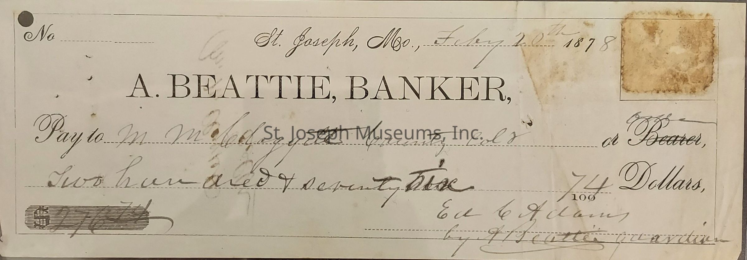 The image shows a vintage check from St. Joseph, Missouri, dated February 20, 1878, issued by A. Beattie, Banker. Written to M.W. Caughey, or Bearer, it authorizes the payment of two hundred seventy-five dollars. The monetary amount is written in both text and numerals, with signatures at the bottom, presumably to validate it. The check bears marks indicative of its age, including a brown stain on the top right corner and a hole-punched section near the top left, perhaps used for bookkeeping or filing purposes. The cursive handwriting, which was more commonly utilized in formal and financial documents of that era, adds an air of authenticity and historical value to the artifact.