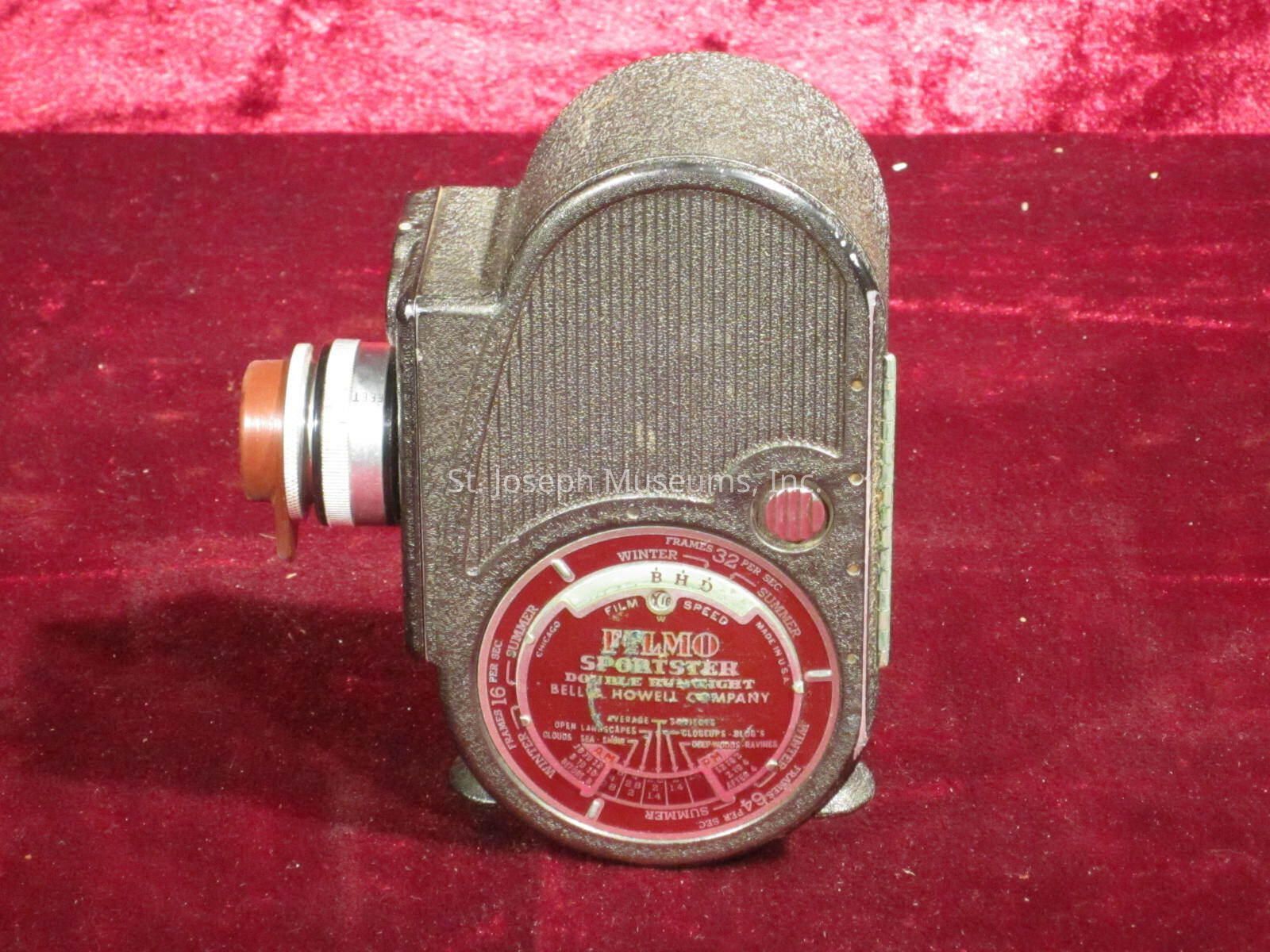 The image displays a vintage Cine-Kodak camera with a textured metallic body and a conspicuous red dial on its side, set against a vibrant red velvet background. The camera features a classic design with a prominent lens to the left, indicative of early 20th-century photographic equipment. Its rounded edges and detailed engravings emphasize its retro aesthetic and engineering precision. This collectible piece likely hails from an era when such cameras were pivotal in capturing home movies, reflecting both technological advancement and cultural significance.