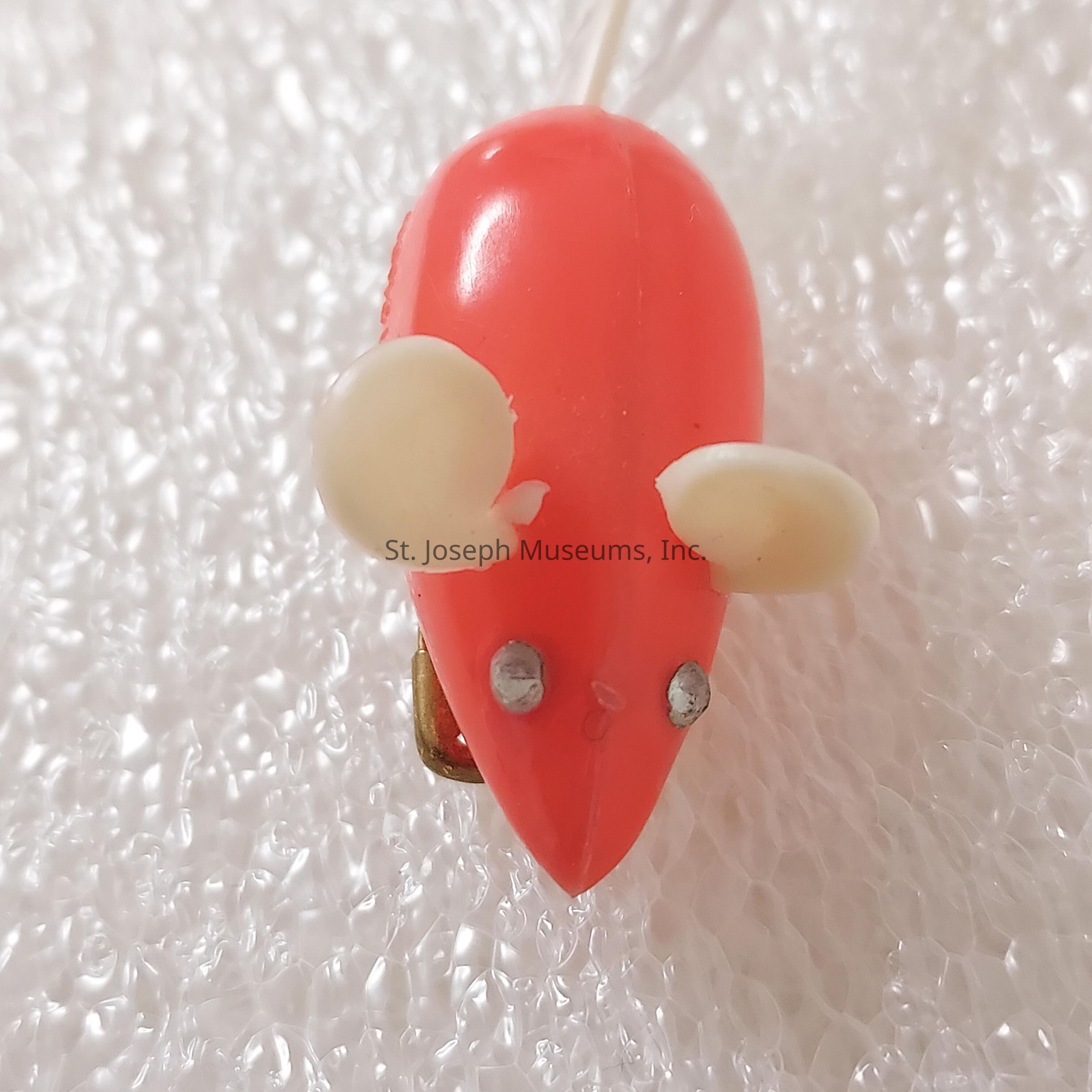 The image depicts a small fishing lure designed to resemble a mouse, featuring a pointed, vibrant red body that stands out prominently against the textured, white background. The lure includes two circular white ears made from a different material attached to the top, completing the mouse-like appearance. Below these ears, two metallic spots serve as eyes, giving the illusion of a face. The shiny texture of the lure reflects light, emphasizing its smooth and glossy surface. Additionally, faint lines are present, adding subtle detailing to the lure's design, possibly representing whiskers or markings. The background consists of numerous small bubbles or particles, resembling foam, creating a contrasting texture that highlights the lure's distinct features. Overall, the image captures the bright, colorful, and imaginative design of a cleverly crafted fishing accessory intended to attract fish by mimicking a small rodent, utilizing vibrant color and playful aesthetics.