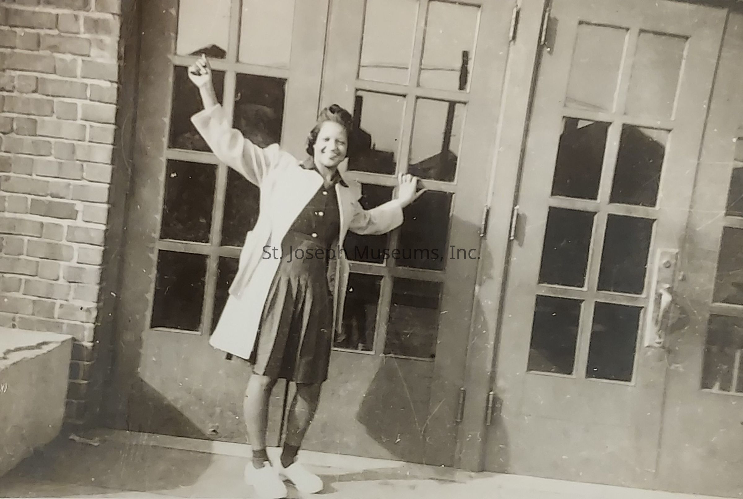 In a vintage black and white photograph, a young woman stands joyfully in front of a large set of glass-paneled double doors, her arms playfully stretched out to each side, and holding an object in one hand, while she wears a dark dress beneath a light-colored coat, complemented by white ankle socks and shoes; the shadowy reflections in the doors and the brick wall at her side hint at a moment captured in a bygone era, evoking a sense of nostalgia with the overall composition and attire, suggesting a setting perhaps outside a school or significant building where moments of joy and anticipation are often felt and remembered.