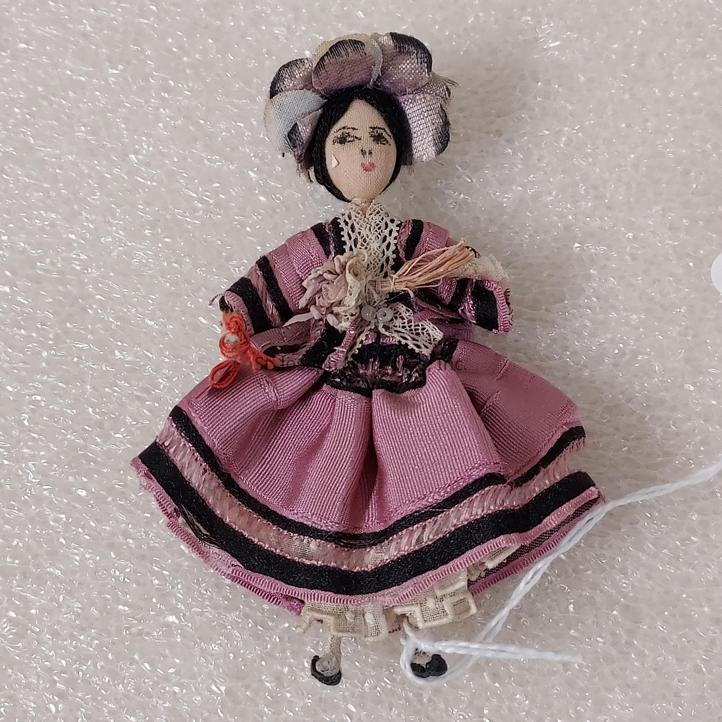 The image depicts a small, handcrafted doll dressed in a vintage-style outfit primarily consisting of pink fabric with black stripes. The doll has a detailed, embroidered face with delicate features such as eyes, a small nose, and red lips. On its head, the doll wears a hat adorned with ruffled fabric in varying shades of purple and white, adding a touch of elegance. It appears to be holding a small piece of red thread or ribbon in one hand. The doll's dress features intricate lace and ribbon detailing, with a cinched waist and a voluminous skirt. The background consists of a textured, off-white surface, which contrasts with the brighter shades of the doll, bringing attention to its handcrafted details. The overall presentation suggests a sense of nostalgia and craftsmanship, highlighting the artistic effort and creativity involved in its creation.