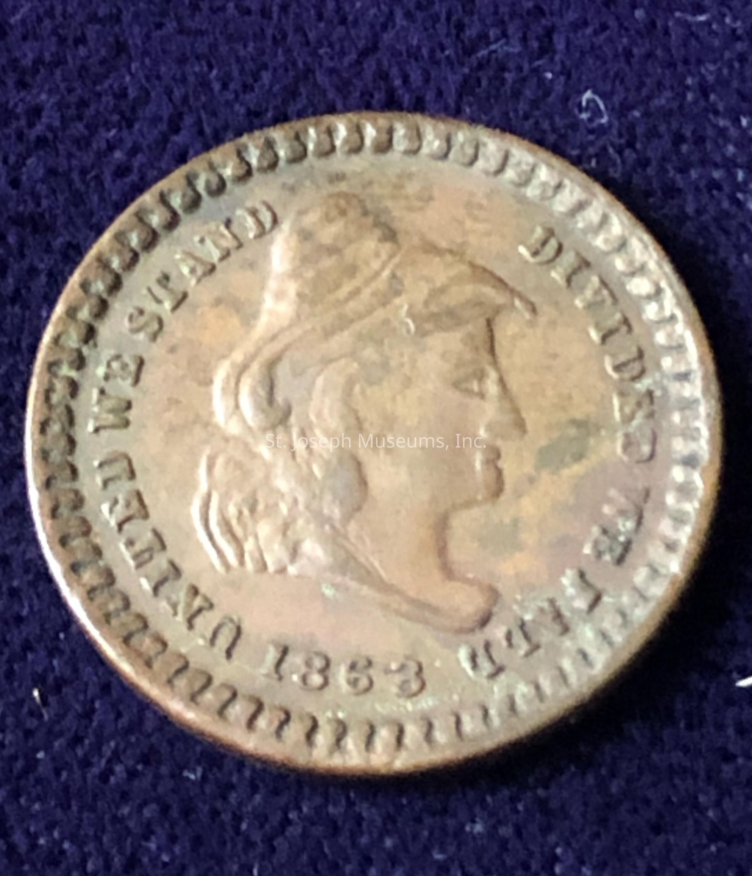 T The coin features a detailed profile of a figure, possibly a woman, wearing a headpiece, situated at the center. Around the edges of the coin, there are inscriptions, though they appear slightly worn due to the age and usage of the coin. The date "1863" is clearly visible near the bottom, and a phrase, "UNITED WE STAND DIVIDED WE FALL," can be discerned circling the perimeter. The coin's surface shows signs of aging, with a patina that adds to its historical character.