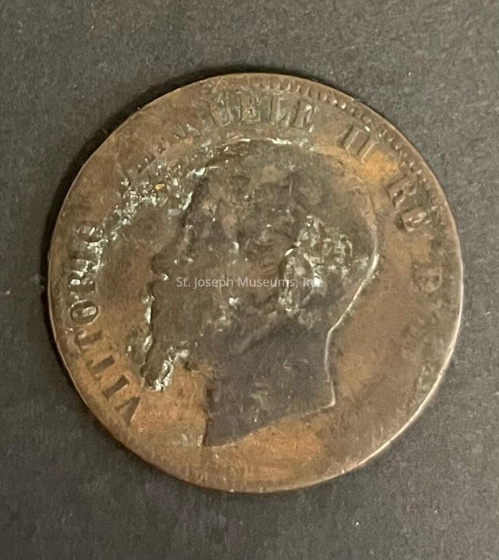 The image depicts an old, worn coin with a profile of a male figure embossed on its surface, surrounded by inscriptions including "VITTORIO EMMANUELE II RE," suggesting its historic value and connection to Italian history, as it likely represents King Victor Emmanuel II of Italy. The coin's surface appears tarnished, indicating its age and potential exposure to the elements over time, which adds to its antique charm. The background is a plain, dark surface, allowing the coin to be the focal point of the image, highlighting its intricate detailing despite its worn condition.