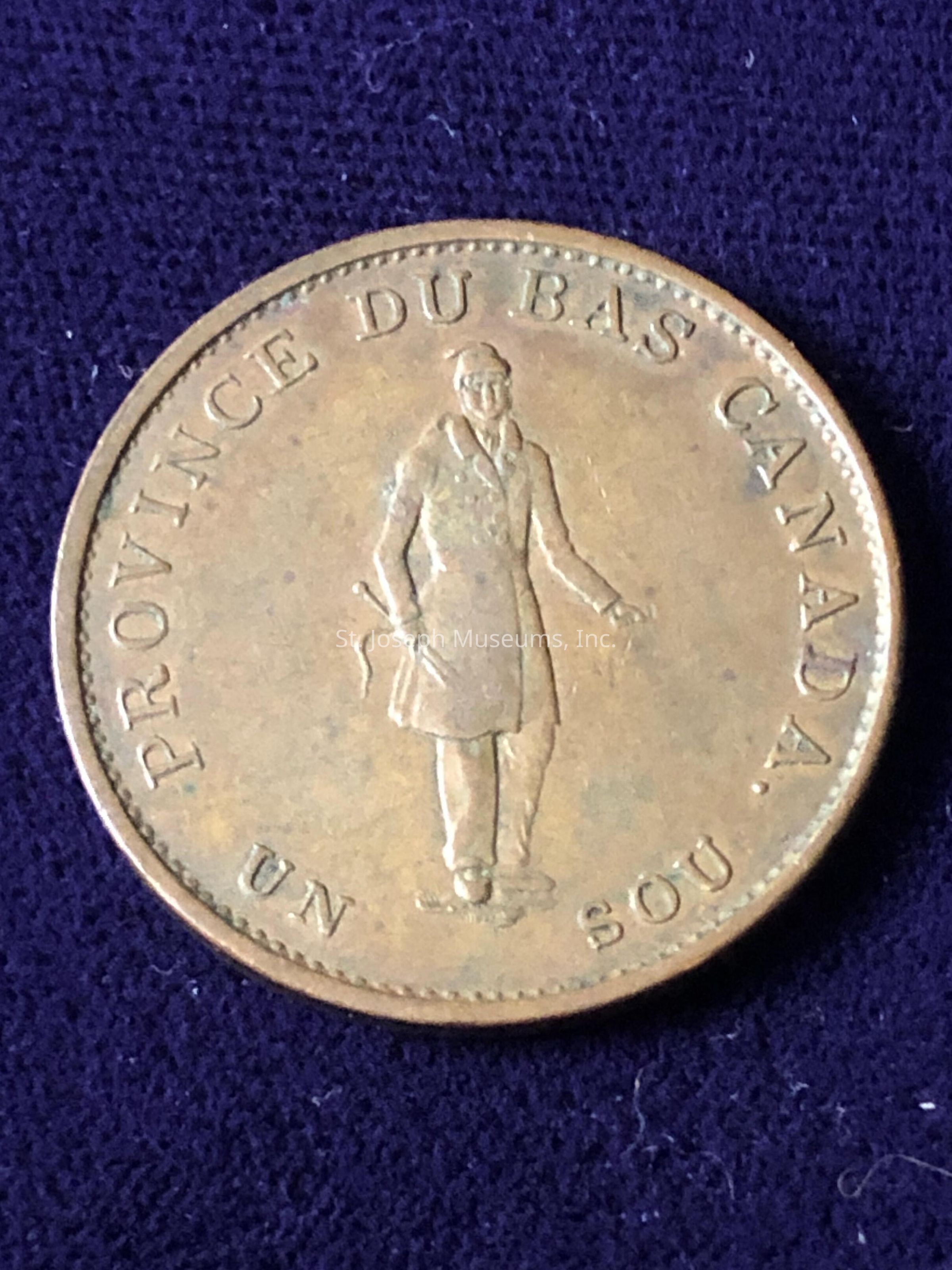 The image depicts an antique coin with the inscription "PROVINCE DU BAS CANADA" and "UN SOU" along its edge, indicating its historical origin from Lower Canada. At the center of the coin, there is an engraved figure of a person dressed in a coat, holding objects in both hands, against a textured surface. This coin, made from what appears to be a bronze or copper alloy, presents a vintage appearance, with visible signs of age and wear, such as tarnish and minor surface imperfections. The background features a dark blue or purple fabric, providing a contrast that highlights the coin's details and coloration. This piece likely serves as a collectible item, offering insights into the historical currency and socio-economic context of 19th-century Canada, reflecting the design and minting techniques of that era. The coin's detailed craftsmanship, including the finely engraved text and figure, suggests a high level of skill in its creation, typical of currency from that period, when such coins were used in