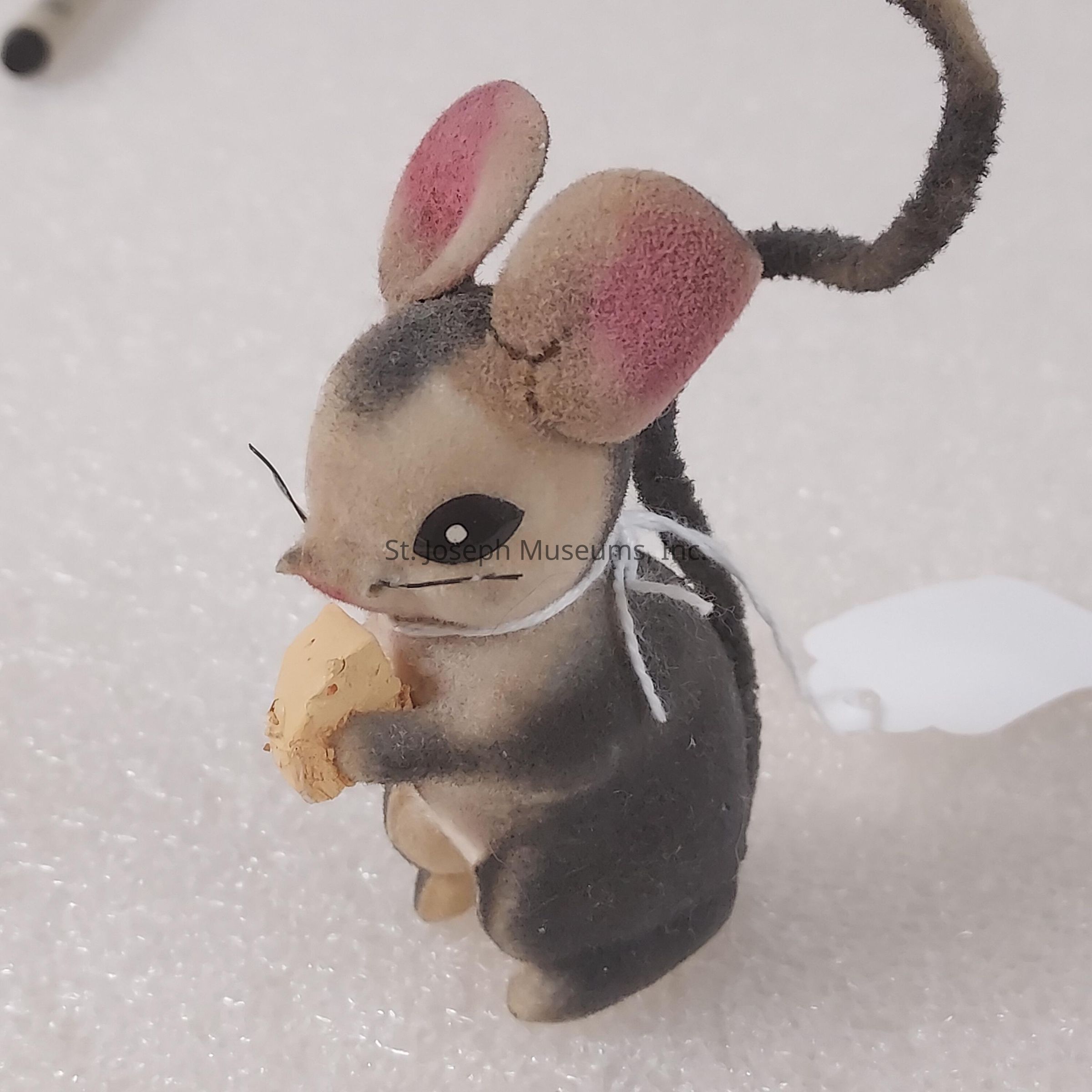 The image displays a small, intricately designed plush mouse figurine with a soft gray and brown fur texture, large pink inner ears, and a long, curled tail. The mouse is standing upright, holding a small piece of yellow cheese in its front paws. It has a black-eye expression, giving it a playful and curious demeanor. A white string tied around its neck suggests that it might be a decorative piece or ornament. The background is a plain, textured surface, allowing the detailed features of the mouse figurine to stand out distinctly.