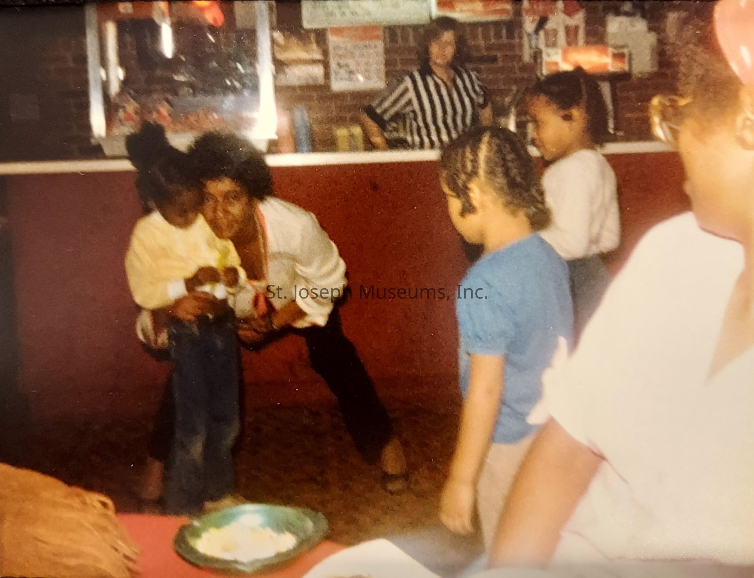 In the image, a group of individuals is gathered in a warmly lit, cozy setting that appears to be an informal social gathering or celebration. A person, with curly hair and clad in a light-colored shirt, is bending down to share a joyful moment with a young child dressed in a yellow top, holding a small object. Nearby, two other children, one wearing a white shirt and another in a blue shirt, are playfully engaged in the scene. In the foreground, an adult sits, partially visible, while a person in a referee-style shirt stands in the background against a brick wall, overseeing the lively atmosphere. On a nearby table, a bowl with some remaining food reflects signs of a pleasant dining experience.