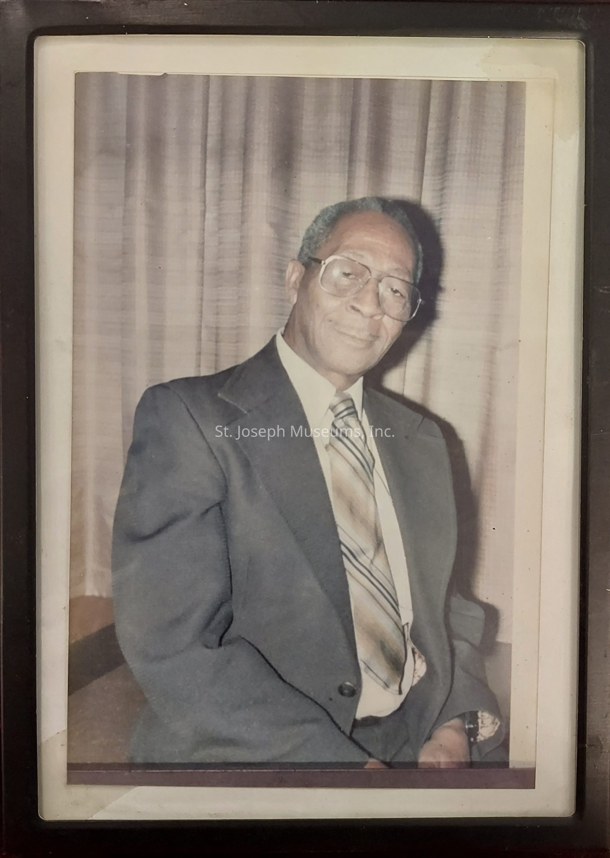 The image portrays an elderly man seated gracefully in front of a plain, slightly textured curtain that suggests an indoor setting. He is dressed in a formal, dark suit that fits him comfortably, highlighting a white shirt beneath it. A patterned, multi-striped tie elegantly shifts down his chest, with subtle color contrasts, adding a touch of character to his attire. His posture is relaxed; he sits with an air of dignified composure, suggesting a sense of wisdom and experience. His face displays a gentle expression, further accented by the large, clear glasses on his face that rest securely, drawing attention to his thoughtful eyes. His hair, short and neatly styled, appears tinged with the natural progression of age. The combination of his attire, demeanor, and the setting conveys an impression of formality, respect, and timeless elegance, embodying a person whose life experiences speak volumes, even in silence. The photograph is encased in a classic frame, emphasizing its significance and the value it holds.