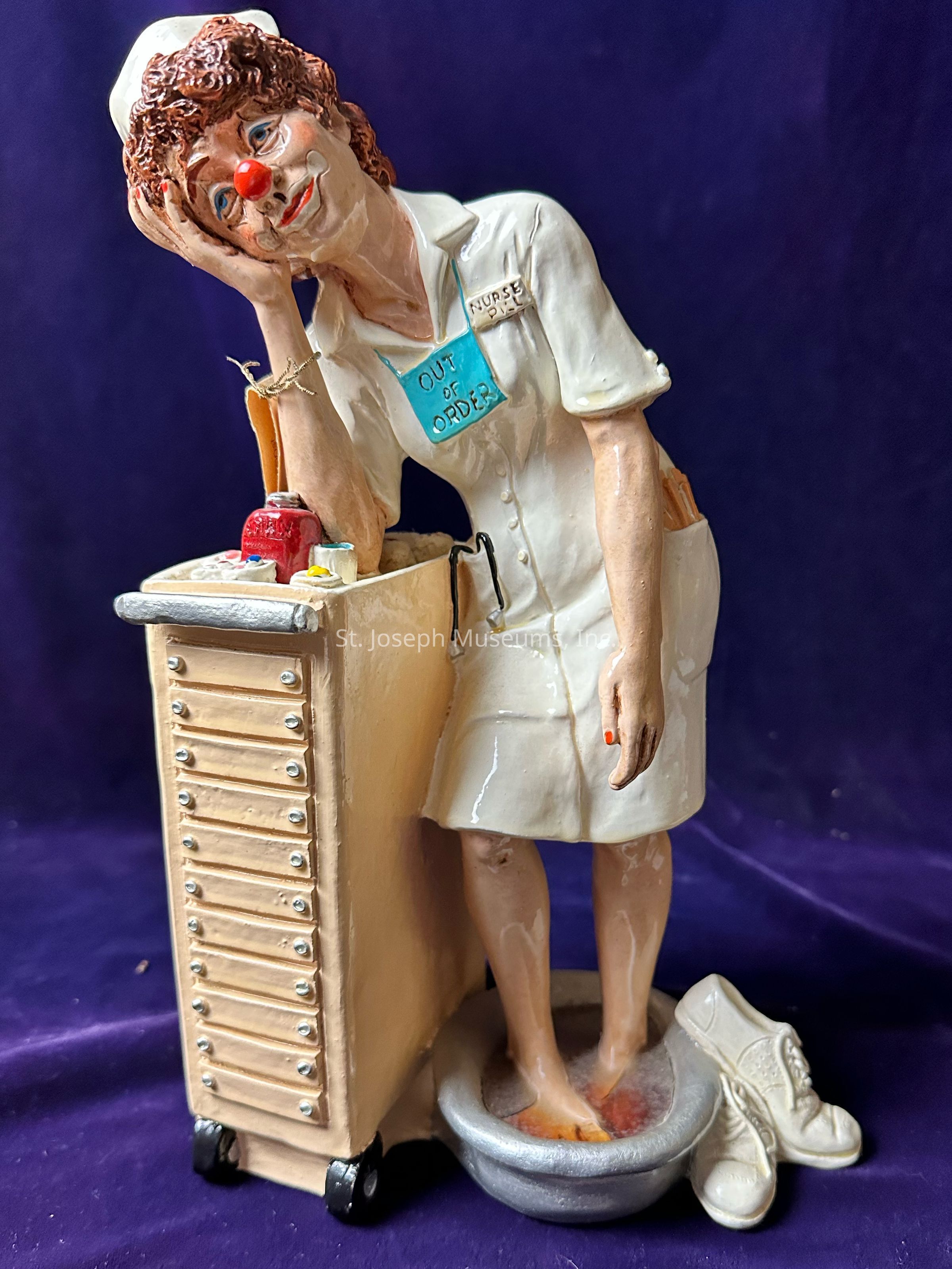 The image depicts a whimsical figurine of a nurse, characterized by a playful expression and wearing a white uniform with a name badge that reads "NURSE," along with a humorous "OUT OF ORDER" sign. Her curly hair peeks out from under her nurse cap, and a red clown nose adds a comical touch to her face. Resting her head on her hand, she leans against a well-detailed medical cart stocked with tiny items. Her bare feet soak in a small foot bath beside her neatly placed shoes. The purple background contrasts with the figurine's details, emphasizing the lighthearted and relaxed atmosphere conveyed by the nurse's pose and features.