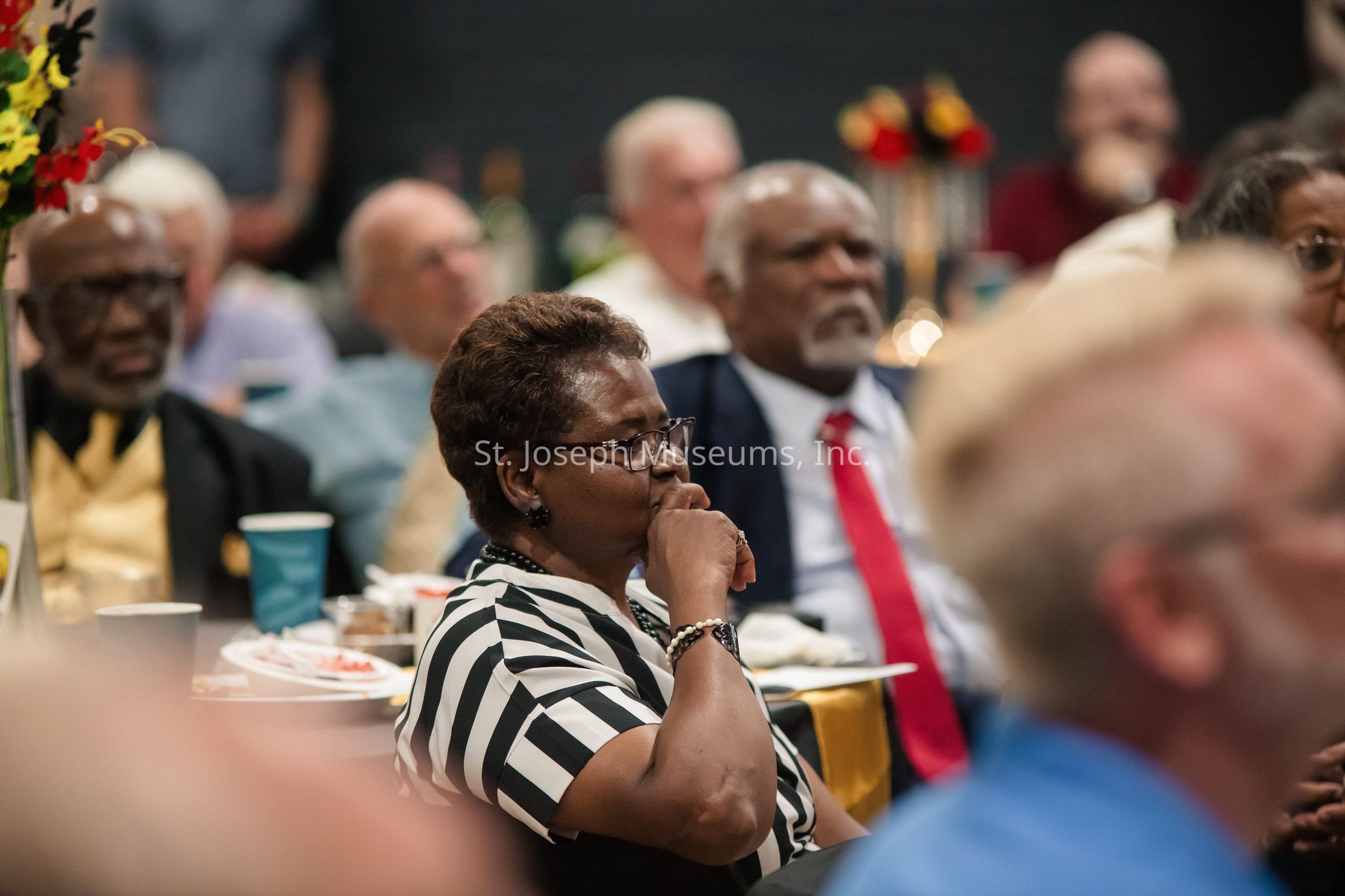 In the image, people are seated at a formal event, paying attention to something out of frame, with a woman in the foreground wearing a striped shirt, glasses, and a thoughtful expression, suggesting she is engaged in the proceedings; she is surrounded by others, mostly older individuals, including men in suits and ties, creating a sense of a gathering that might be a conference, reception, or award ceremony, with neatly arranged tables featuring plates, cups, and decorations, indicating a well-organized event space with a social and professional atmosphere.