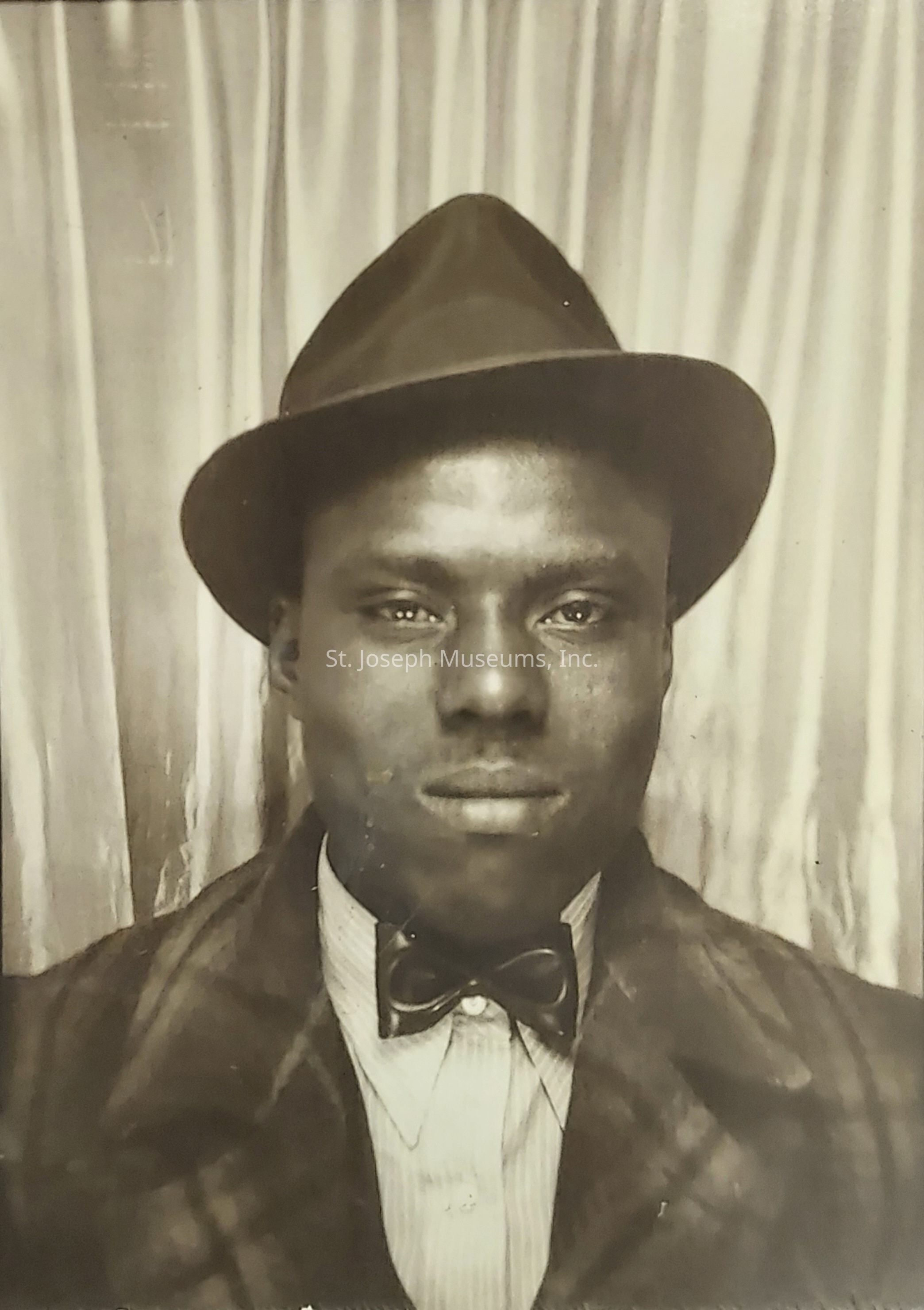 The image is a vintage black and white portrait of an unidentified man, who is looking directly at the camera with a neutral expression, wearing a fedora hat and a plaid coat; under the coat, there is a collared shirt paired with a bow tie, indicating a formal attire typical of a past era, and the background consists of simple, vertical curtains which are slightly out of focus, suggesting that the photograph might have been taken in a studio or a photo booth; the overall composition of the image, including the clothing, style, and monochrome tone, evokes a sense of timelessness and historical context, highlighting the fashion and photography style of the time, while the man's direct gaze and poised expression convey a sense of confidence or seriousness, making the portrait intriguing and inviting viewers to ponder the story or life of the person captured in this moment.