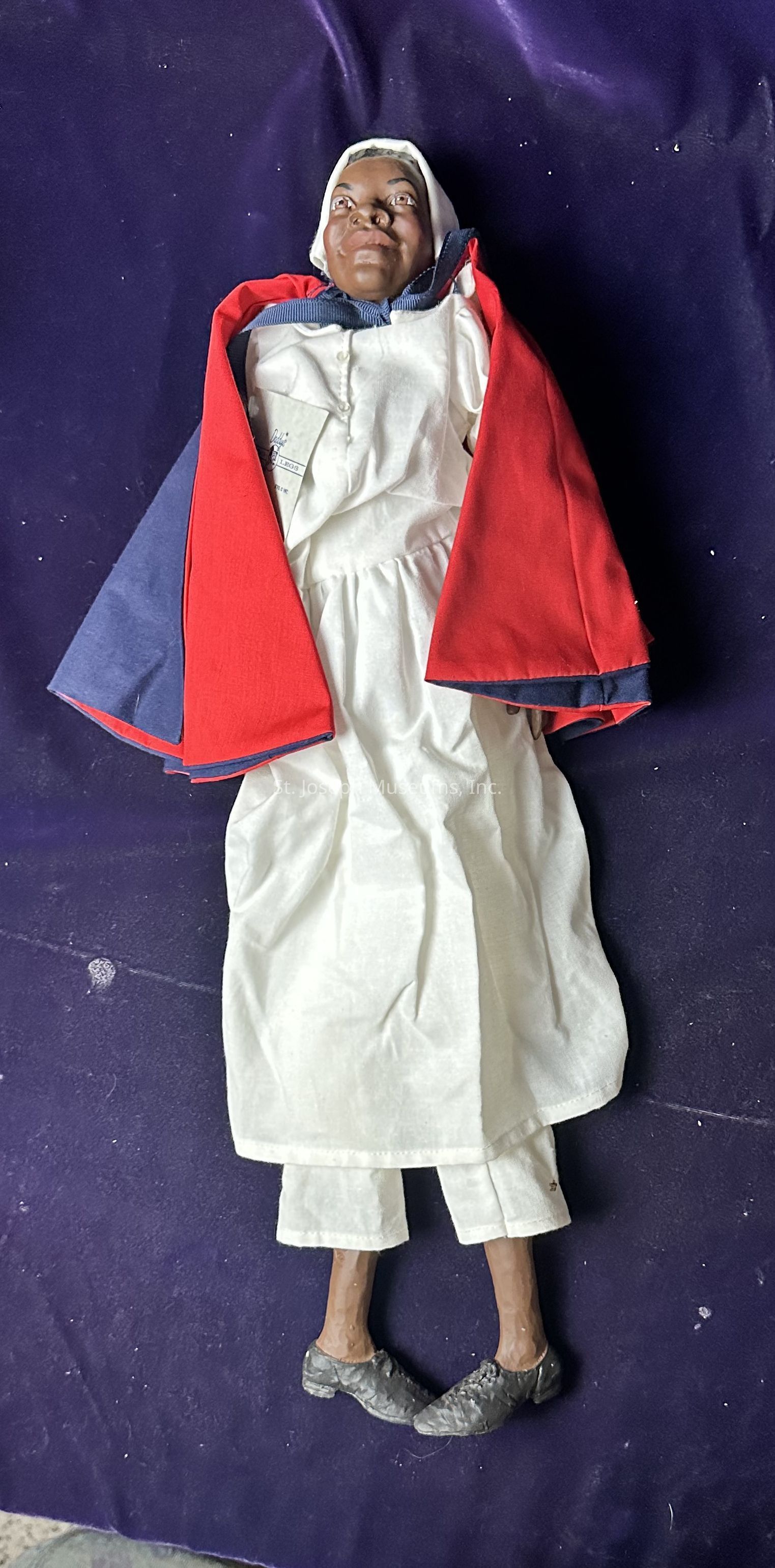 The image depicts a doll lying on a deep purple surface, wearing a white dress and a matching headscarf. Over the dress, a vibrant red and blue cloak is draped around the doll’s shoulders. The doll is also wearing black shoes and has distinct facial features, with a serene expression. The outfit is detailed, with visible stitching and button accents on the garment, suggesting it is carefully crafted. The lighting highlights the textures of the clothing and surface, providing a clear view of the doll's attire and the soft, smooth fabric of the backdrop, creating a visually appealing composition.