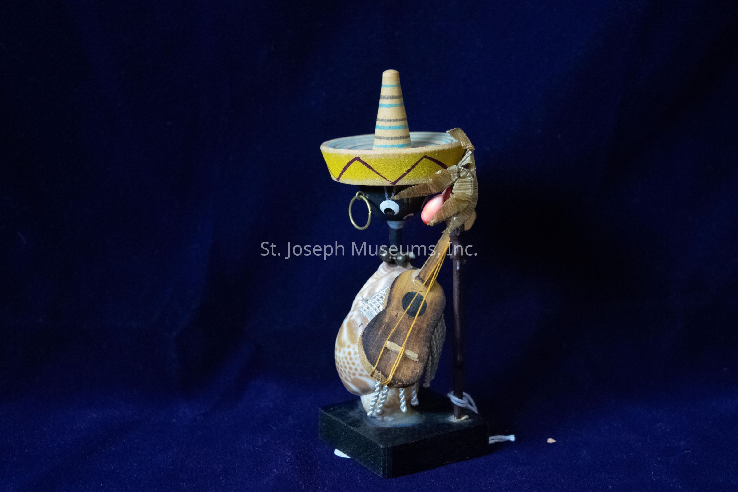 The image shows a whimsical figurine crafted primarily from a shell, which forms the body of the character. This quirky figure is adorned with a colorful sombrero, adding an element of cultural flair to its appearance. The character has comical, googly eyes that give it a playful and animated expression. An earring hangs from what appears to be one of its ears, contributing to its quirky personality. A small wooden guitar is slung over the shell, suggesting that this character might be a music enthusiast or musician. The figurine stands on a sturdy black base, and the background is a deep, rich blue, providing a vibrant contrast that allows the colors and details of the figurine to stand out. This imaginative piece of art seems like a delightful piece of decoration, blending humor with creative craftsmanship.