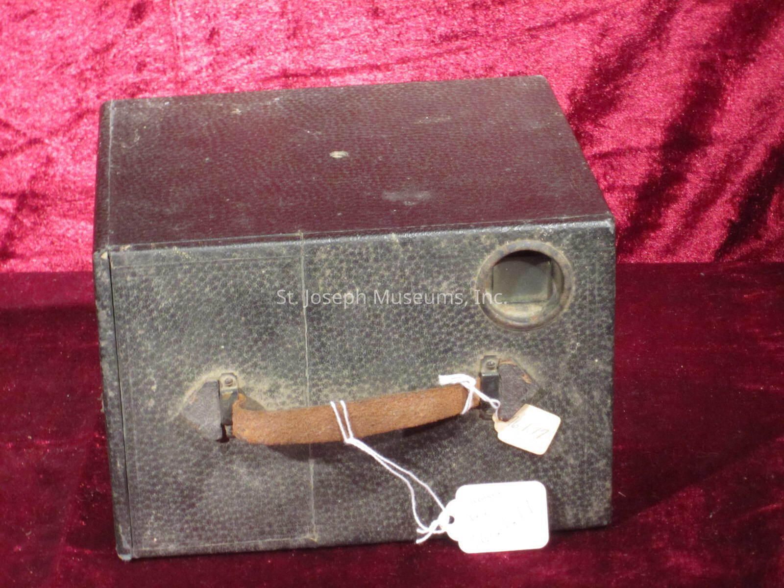 The image depicts an old, vintage box camera with a textured, dark outer casing that appears worn with age. Its simple, rectangular shape is complemented by a small circular lens opening on one side, while a leather strap is affixed to the front, suggesting portability. Attached to the strap are two white tags, hinting that the item might be part of a collection or sale. The camera is set against a rich, burgundy fabric background, which adds an elegant contrast to the rustic look of the item. The overall appearance suggests a historical artifact, possibly from the early 20th century, used for basic photography.