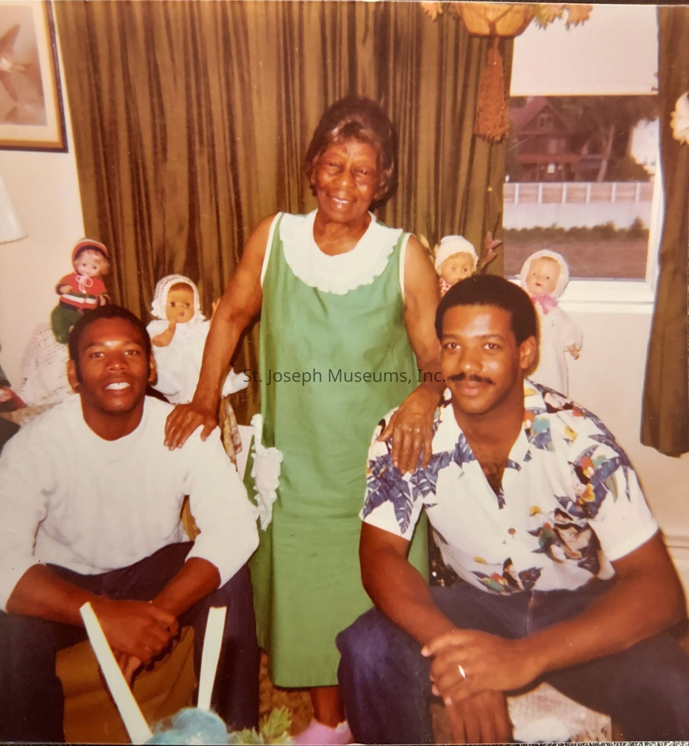 The image shows three people inside a warmly decorated room. An elderly woman, standing in the center and smiling, is dressed in a green dress with a white collar. Her hands rest affectionately on the shoulders of two men sitting beside her. The man on the left is wearing a white sweater, while the man on the right sports a colorful short-sleeved shirt. Numerous dolls with various outfits and hats are neatly arranged on a table behind them, creating a cheerful and nostalgic atmosphere. A window in the background reveals a glimpse of houses and a white fence outside, while thick, dark green curtains frame the scene inside, indicating a cozy family environment. The warm colors and casual posture of those in the photograph suggest a happy, heartfelt moment being captured, possibly a family gathering or celebration.