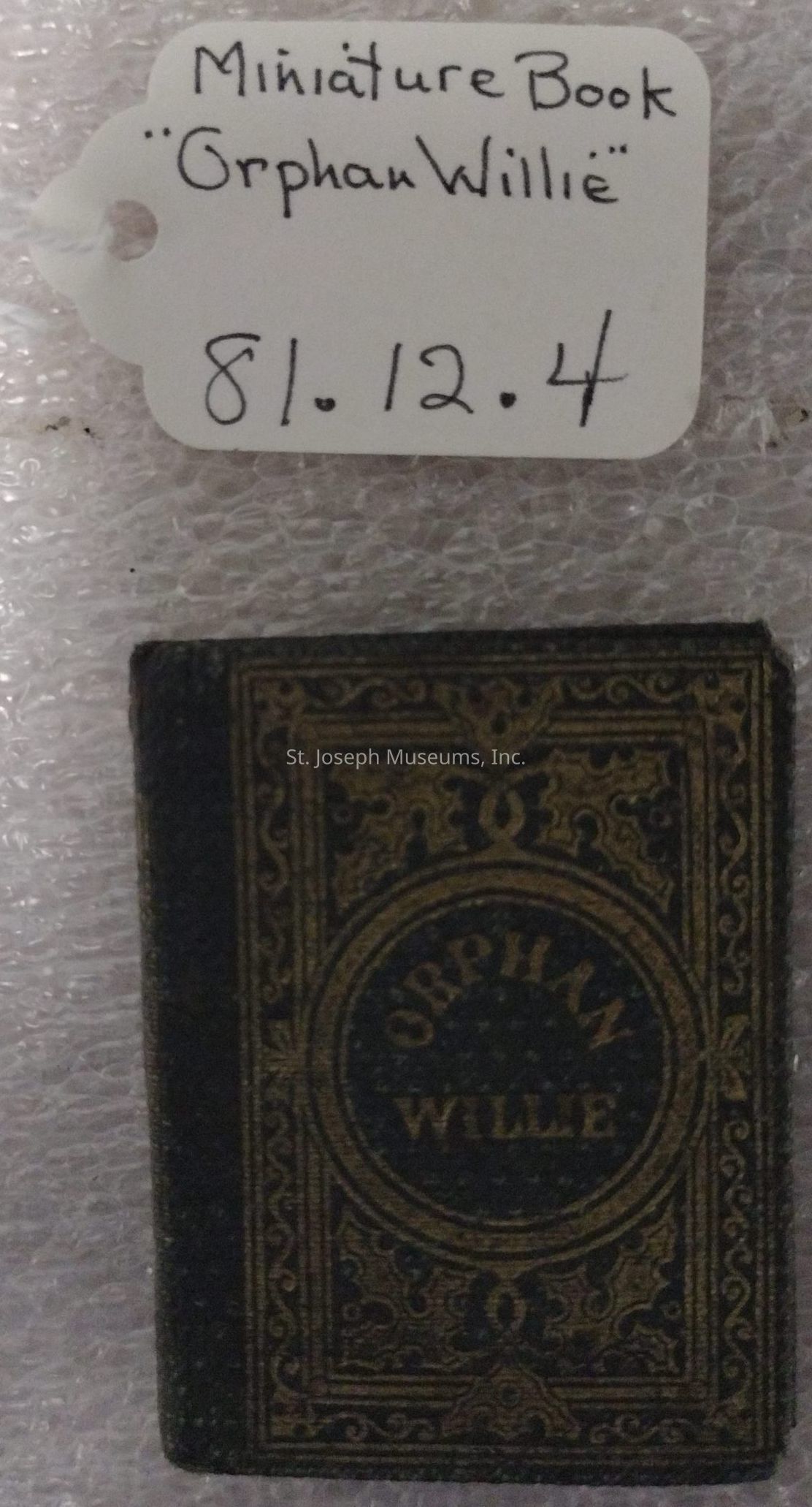 The image shows an intricate miniature book titled "Orphan Willie," with a decorative cover design featuring ornate patterns, likely in gold against a dark background. Above the book, a handwritten tag provides information, describing the item as a "Miniature Book" with the title "Orphan Willie" and an inventory number "81.12.4." The book and its tag are on a textured surface, possibly bubble wrap, suggesting careful handling or storage. The detailed design and small size imply that this book might have been a collector's item or part of a specific exhibition of rare or unique books, emphasizing its cultural or historical significance.