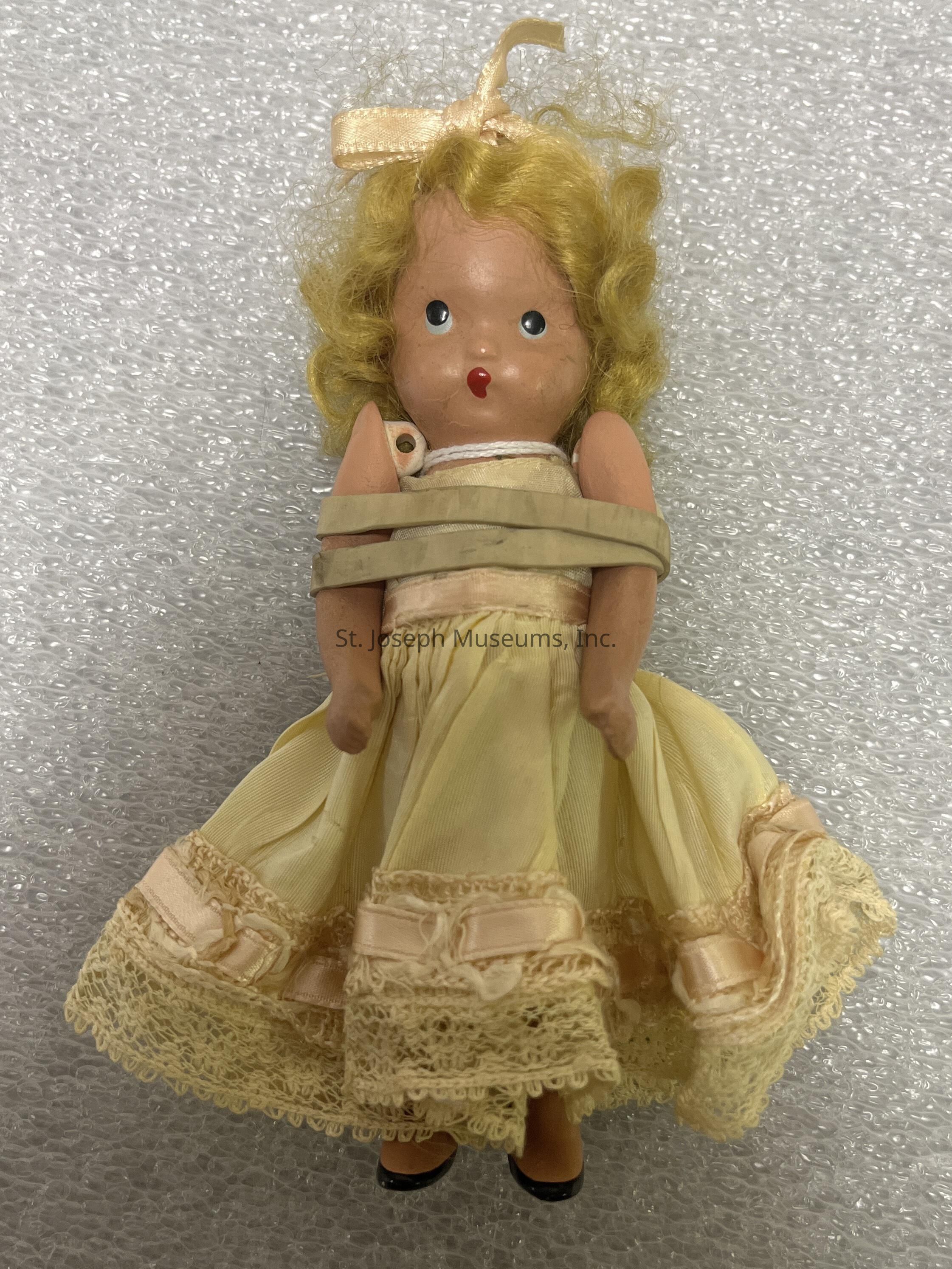 The image shows a vintage doll with curly blonde hair tied with a light-colored ribbon. The doll has painted features, including bright blue eyes and red lips, giving her a classic and timeless expression. She is wearing a cream-colored dress adorned with lace and ribbon details at the hem, providing an elegant and delicate appearance. The dress seems to be made from a soft fabric that complements the doll's overall vintage aesthetic. Her arms are positioned by her sides, and she wears small black shoes. The doll is set against a textured white background that appears to be bubble wrap, which adds depth to the image without detracting from the doll's charming features. This doll likely holds nostalgic value and could be a collectible item, reflecting an era of craftsmanship and attention to detail in toy design. The entire composition of the image highlights the intricacy and delicate beauty of the doll, captivating viewers with its representation of vintage charm.