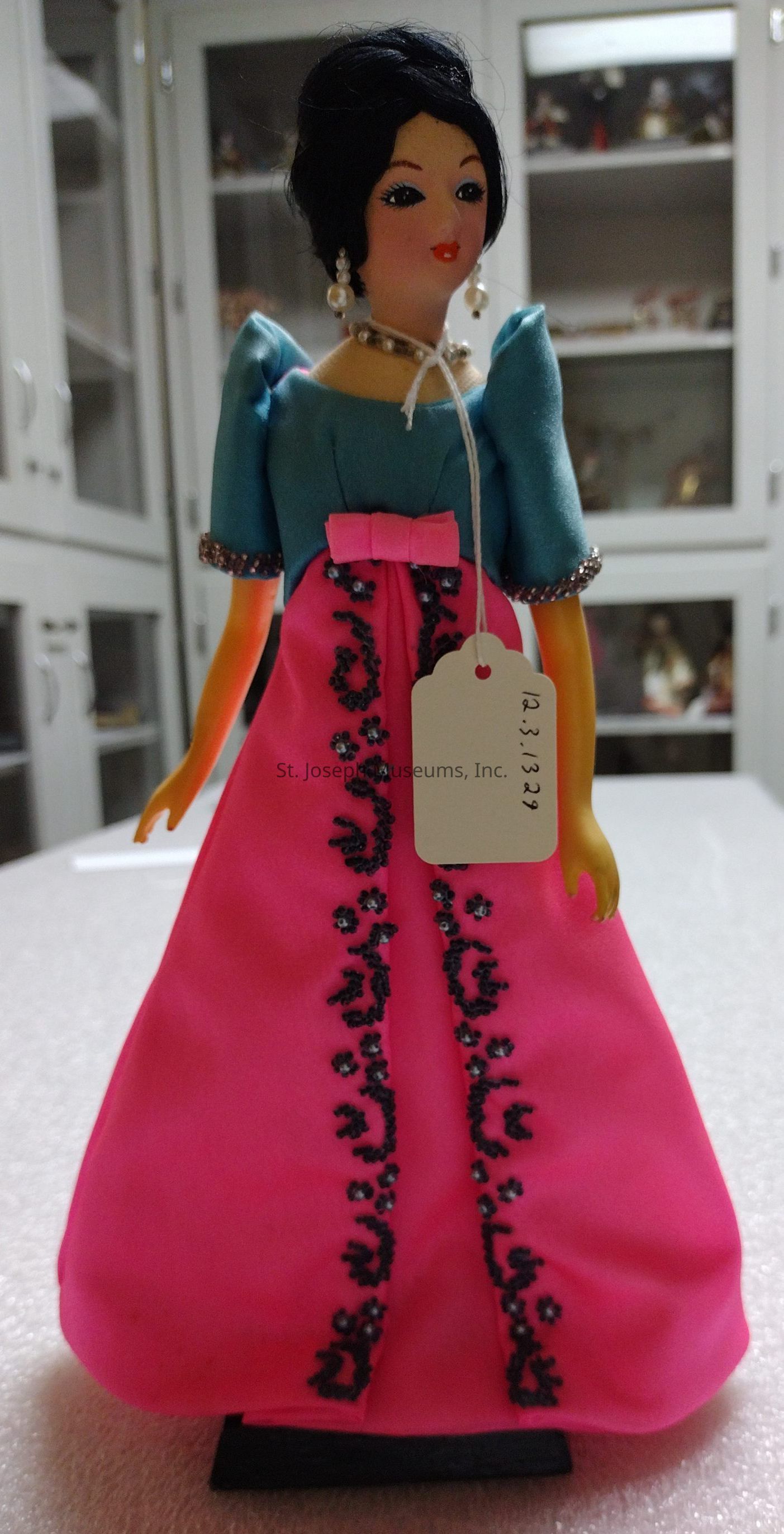 The image shows a detailed doll dressed in a vibrant blue and pink gown, adorned with intricate black embroidery and a pink bow at the waist. The doll features an elegant hairstyle and wears pearl earrings and a beaded necklace, adding elegance to its appearance. A tag with the number "12.3.1329" is attached to the dress, indicating it might be part of a collection or exhibition. The background reveals a cabinet filled with various items, suggesting a display or museum setting. The overall composition highlights the craftsmanship and ornate styling of the doll's attire.