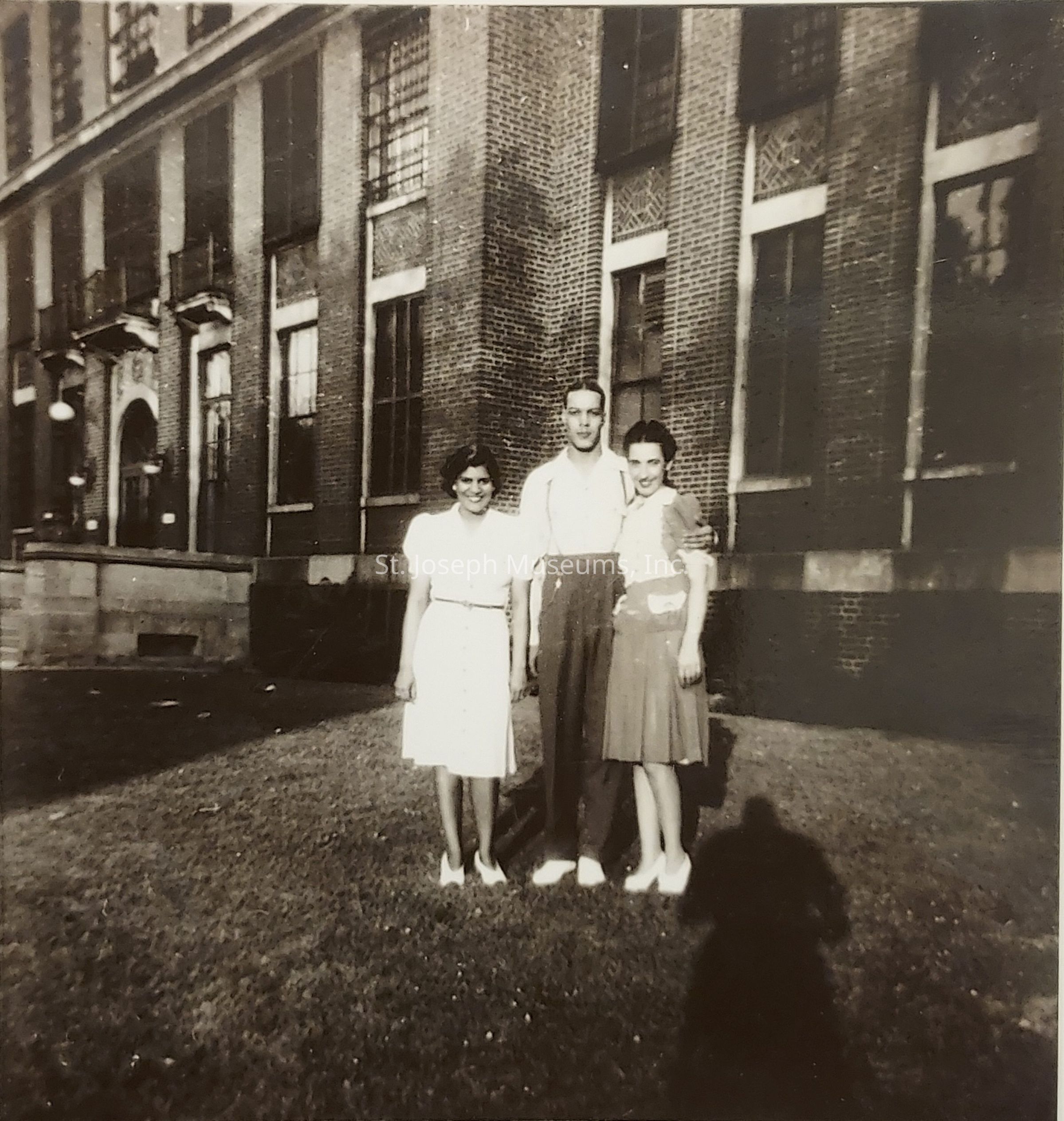 In the black and white photograph, three individuals stand closely together in front of a large brick building with tall windows and intricate latticework, suggesting it might be an educational or institutional structure. Positioned on a neatly trimmed lawn, the group consists of a man flanked by two women, all dressed in mid-20th century attire. The woman on the left wears a light dress with short sleeves and a belt, while the woman on the right, with her arm around the man, is in a slightly darker dress. The man, standing taller between them, wears high-waisted pants and a light shirt. Their expressions convey a sense of camaraderie and relaxation, and a shadow cast on the grass, possibly of the photographer, adds depth to the scene, lending an intimate touch to this seemingly candid moment captured in time, evocative of personal histories and the era's style.