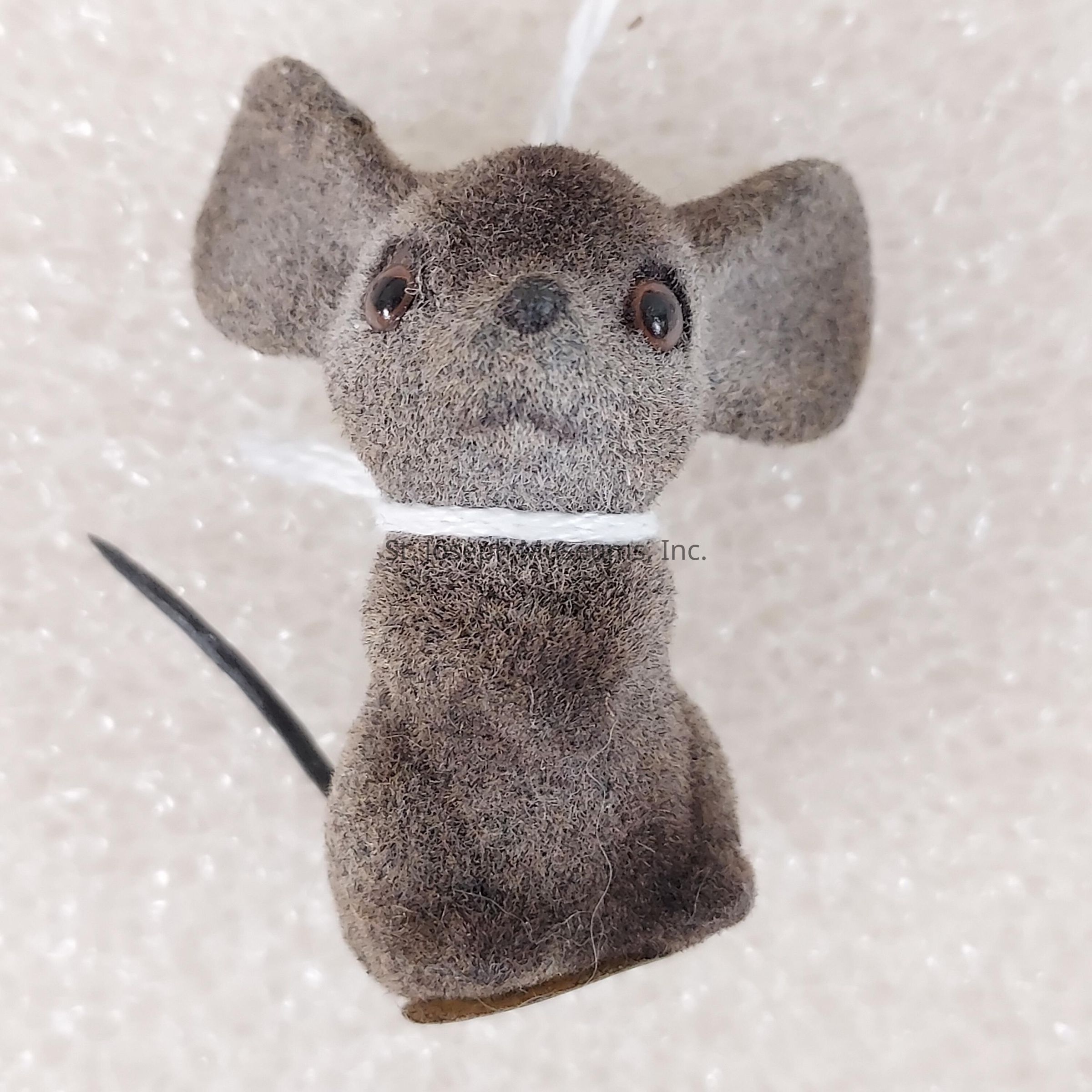 The image shows a small, plush toy resembling a mouse with large ears and a slightly upturned nose, featuring a fuzzy texture and subtle gray color; the toy is tied around the neck with a white string, and its black tail curves slightly to one side, adding balance to its adorable and whimsical appearance against a softly textured background that accentuates its features and emphasizes the cute, handcrafted design.