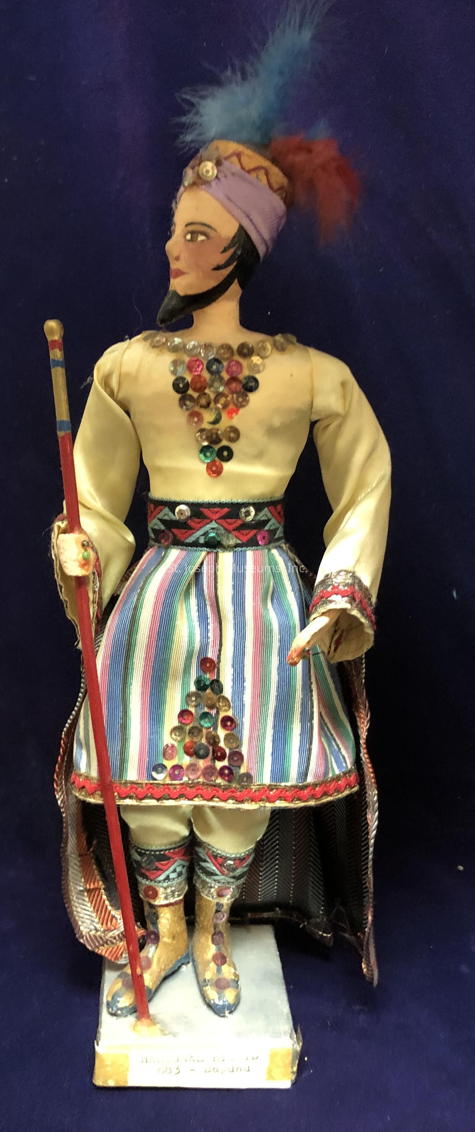 The image features a detailed figurine dressed in traditional attire, set against a deep blue background. The figure wears a hat adorned with colorful feathers in red and blue, complementing the ornate headdress that adds a touch of elegance. The upper garment is decorated with an array of colorful sequins forming a decorative pattern on the chest, which contrasts with the ivory-colored fabric. The lower half showcases a skirt with vertical stripes in vibrant hues, further embellished with sequins. The figure holds a red staff, adding a sense of authority or tradition to its posture. Intricate designs and sequins also adorn the shoes, which rest on a rectangular stand. The figurine exudes a sense of cultural richness and craftsmanship, with attention to detail evident in the choice of colors, patterns, and accessories.