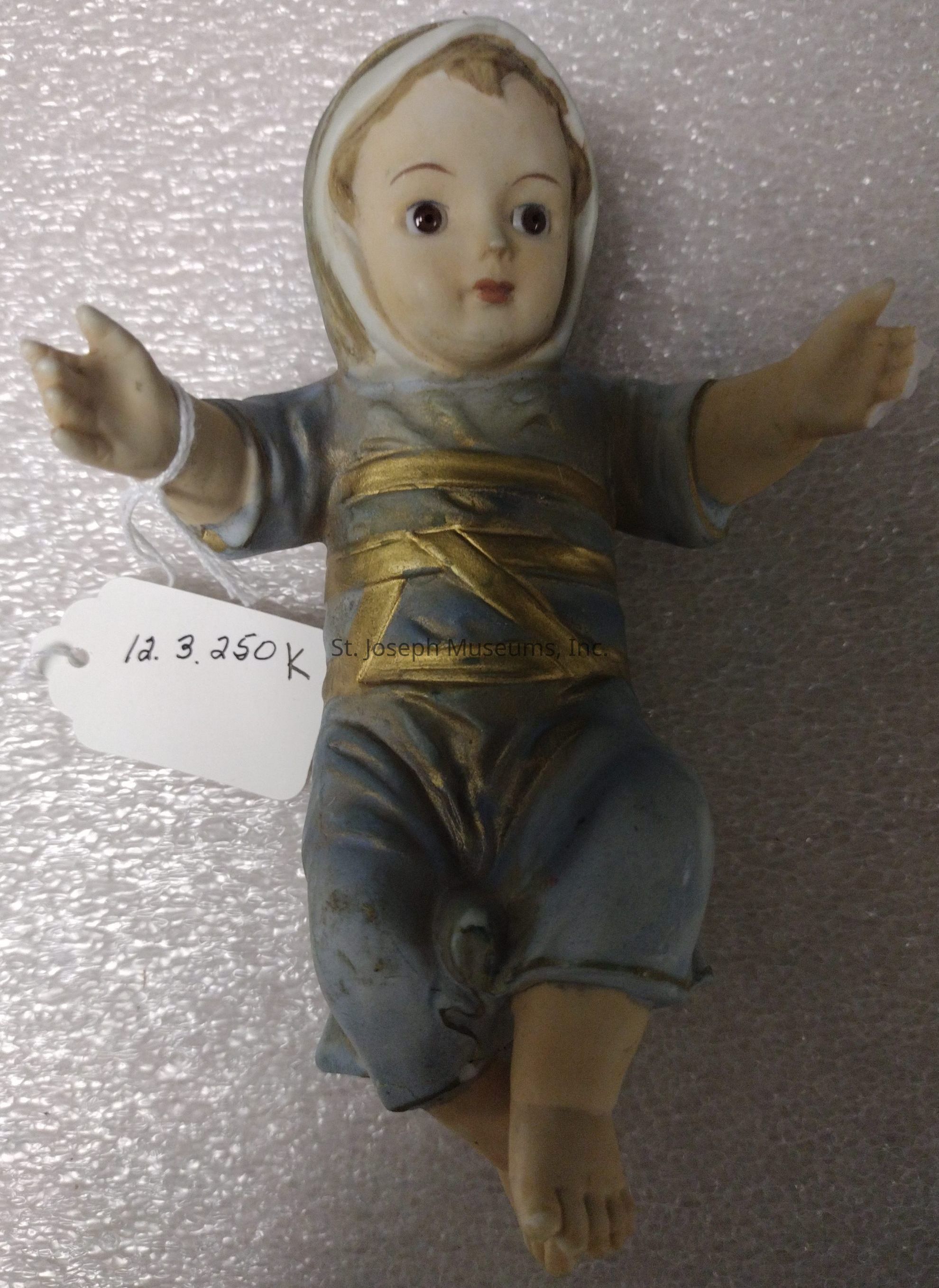 The image depicts a small ceramic figurine of a child, wearing clothing that resembles robes with a hood, which are painted in blue and accented with gold bands across the chest. The figurine has detailed facial features, with wide eyes and a slight smile, giving it a peaceful expression. The child’s arms are open, and the right hand has a loop of string attached to a white tag labeled "12.3.250 K" in handwritten script, suggesting it may be an inventory or pricing marker. The background is a textured, silver-gray surface, which enhances the appearance of the figurine in the foreground.