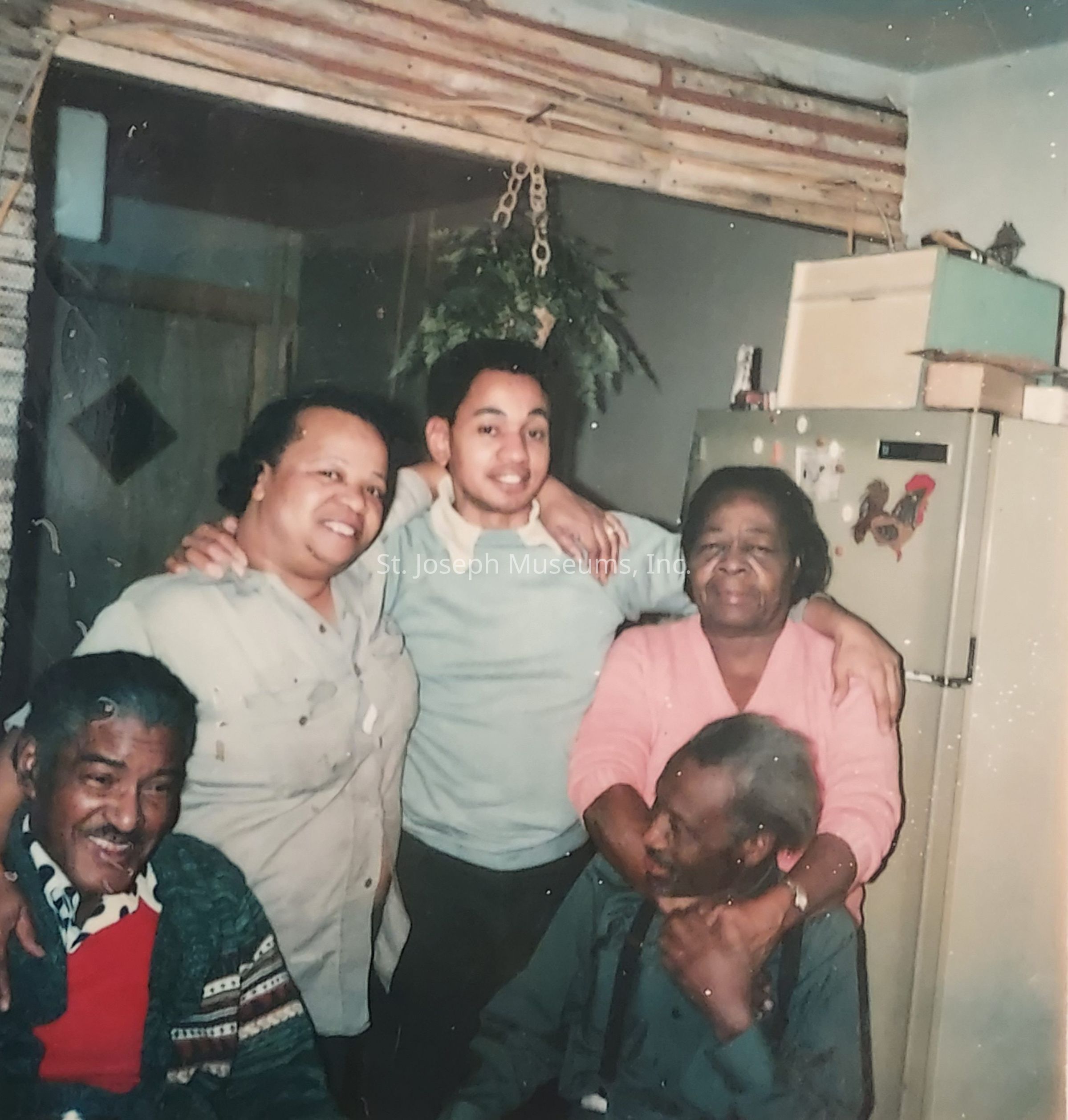 The image captures a warm and intimate family moment inside a cozy room, where five individuals are gathered together, conveying a sense of closeness and affection. There are two women, one with her arms affectionately around a young individual in the center, who's smiling and has his arms around them both. To their right, another woman in a pink sweater is standing next to a seated older man, both sharing a gentle embrace that highlights their bond. On the left, another man is seated, smiling warmly, wearing a patterned sweater. The background features a rustic wooden trim and a hanging pot of greenery, along with a refrigerator adorned with magnets, adding a touch of homely charm to the scene. The overall atmosphere is one of connection, joy, and familial love.