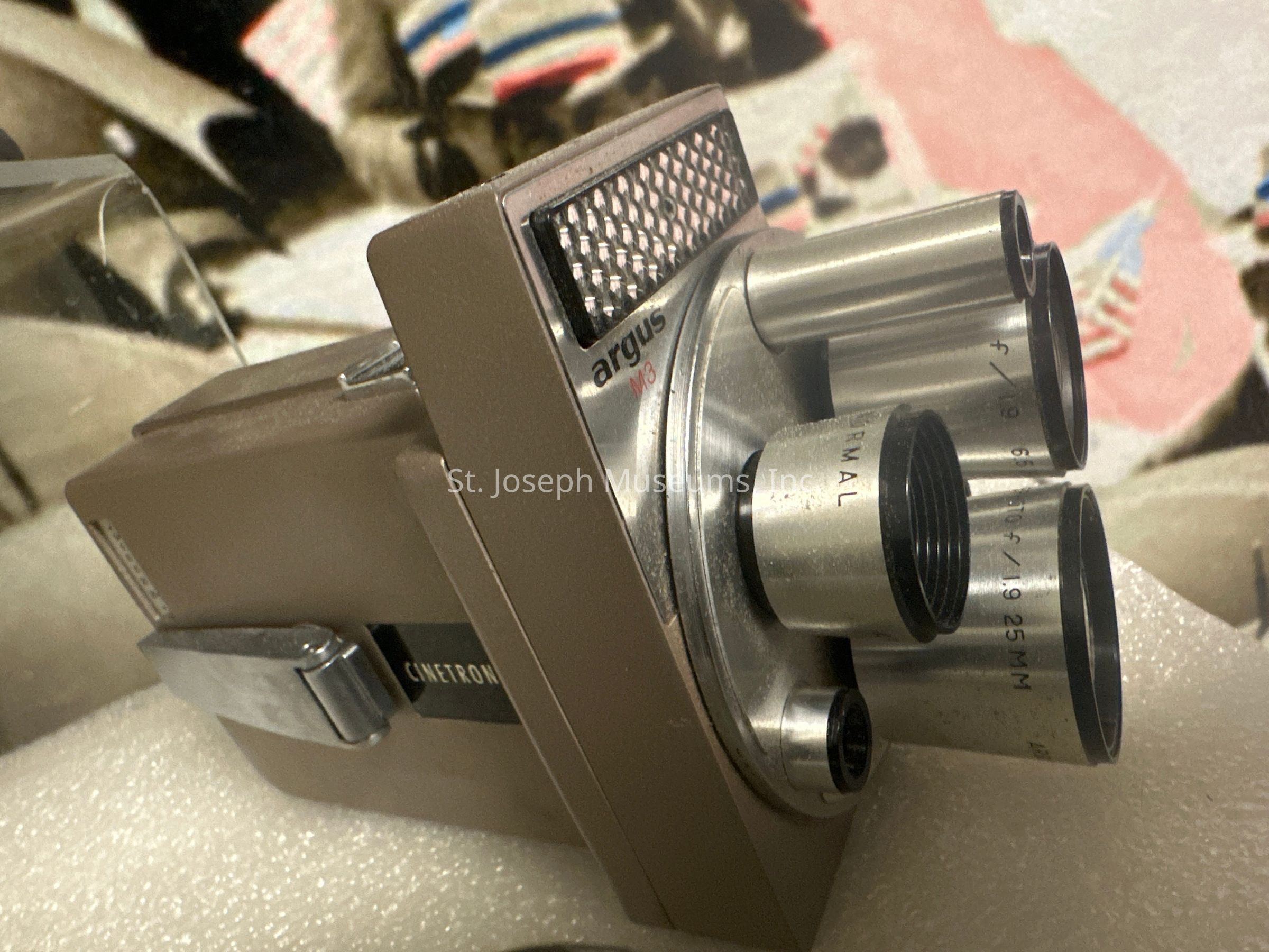 The image depicts a vintage Argus camera featuring an intricate design with multiple lenses protruding from its front. The camera has a rectangular, metallic body with a textured grip area and a prominent viewfinder. The lenses are marked with different focal lengths, indicating its capacity for versatile photography. It is placed against a soft, foam-like surface, providing a clear view of the camera's design details. The background displays a blurred, abstract pattern with soft colors, enhancing the focus on the camera itself. This classic piece of technology showcases the craftsmanship and engineering of its era, adding a nostalgic touch to its aesthetic appeal.