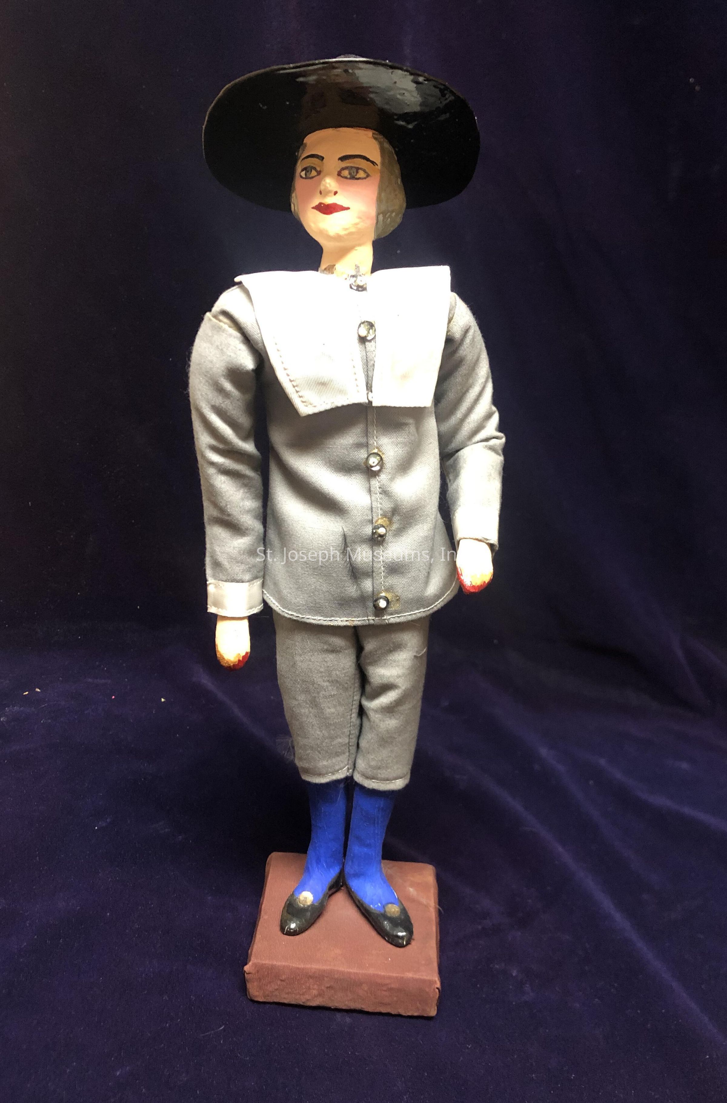 The image features a handcrafted doll standing on a rectangular base, dressed in a grey outfit consisting of a long-sleeved jacket with buttons and matching pants, and a white collar that adds contrast to the attire. The doll is adorned with a wide-brimmed black hat, complementing its painted facial features, including red lips and expressive eyes. Its shoes are fashioned in black with striking blue socks that stand out against the muted tones of the suit. The background is a deep, rich purple, providing an elegant and dark backdrop that highlights the details of the doll's attire and features. The doll's posture, along with the meticulous detailing of its costume, suggests a historical or vintage style, evocative of a bygone era, and reflects careful craftsmanship in its construction, making it a captivating piece of art to observe, likely serving as a decorative or collectible item, displayed for its aesthetic appeal to those with an appreciation for traditional dolls and their intricate detailing.