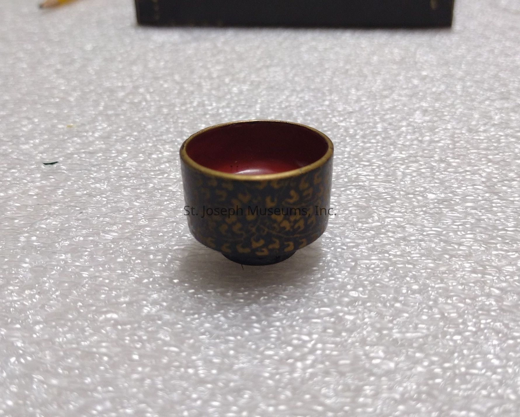 The image features a small, decorative cup set on a textured surface that resembles bubble wrap. The cup is intricately designed, showcasing a dark exterior adorned with a golden, ornate pattern, which imbues it with an element of sophistication and elegance. Its interior is a deep, rich maroon, contrasting beautifully with the exterior design. This combination of colors and patterns suggests that the cup might serve a ceremonial or decorative purpose. The play of light on the bubble wrap surface creates subtle highlights and shadows, adding depth to the overall composition. Additionally, the blurred background slightly hints at other objects, though they are indistinct and unobtrusive, ensuring that the focus remains solely on the cup. The entire scene conveys a sense of delicacy and artistry, making the cup appear as though it could be part of a collection of artisanal or historical artifacts. The intricate detail and craftsmanship point towards a cultural significance, potentially linking the cup to a particular tradition or historical context.