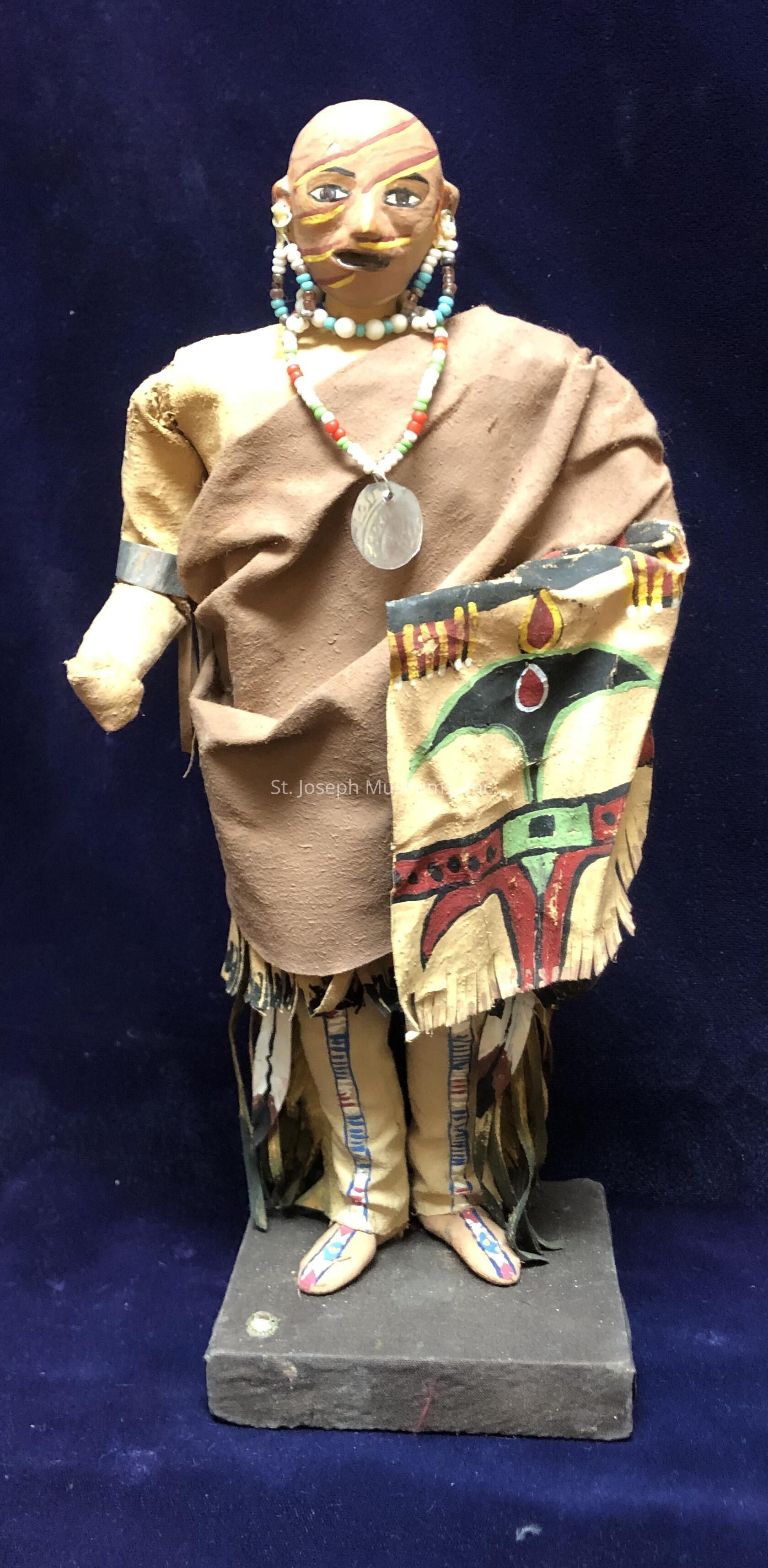 The image shows a handcrafted doll with a distinctly styled head featuring painted details and ornamental beadwork as earrings and a necklace. The doll is dressed in a tan fabric robe with a painted, patterned cloak draped over one shoulder, showcasing colors like red, green, and yellow. It stands on a rectangular base and has additional detailing on its legs, which are adorned with long, vertical fringes and painted designs. The cultural and artistic aspects depicted in this image emphasize intricate craftsmanship and vibrant artistic expression.