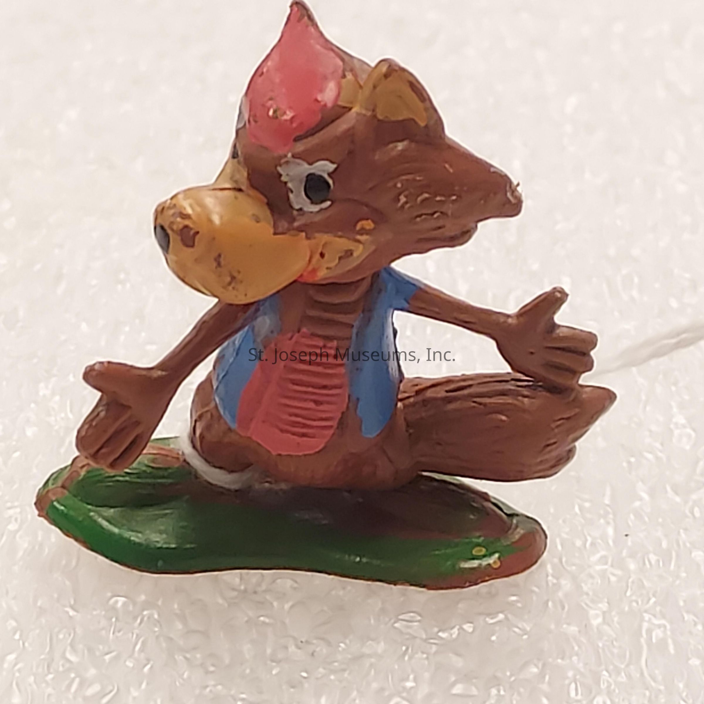 The image depicts a small, colorful, and whimsical figurine of a cartoon-like animal character that resembles a fox or squirrel. The character is painted in bright colors, with a vibrant red tuft on its head, a yellow snout, large round eyes, and a cheerful expression. It is wearing a blue jacket over a red shirt, adding to the playful appearance. The figure's arms are outstretched, and it is seated on a green leaf, which serves as its base, giving the illusion of movement or action. The surface it rests upon appears textured, providing a contrasting backdrop to the lively character.