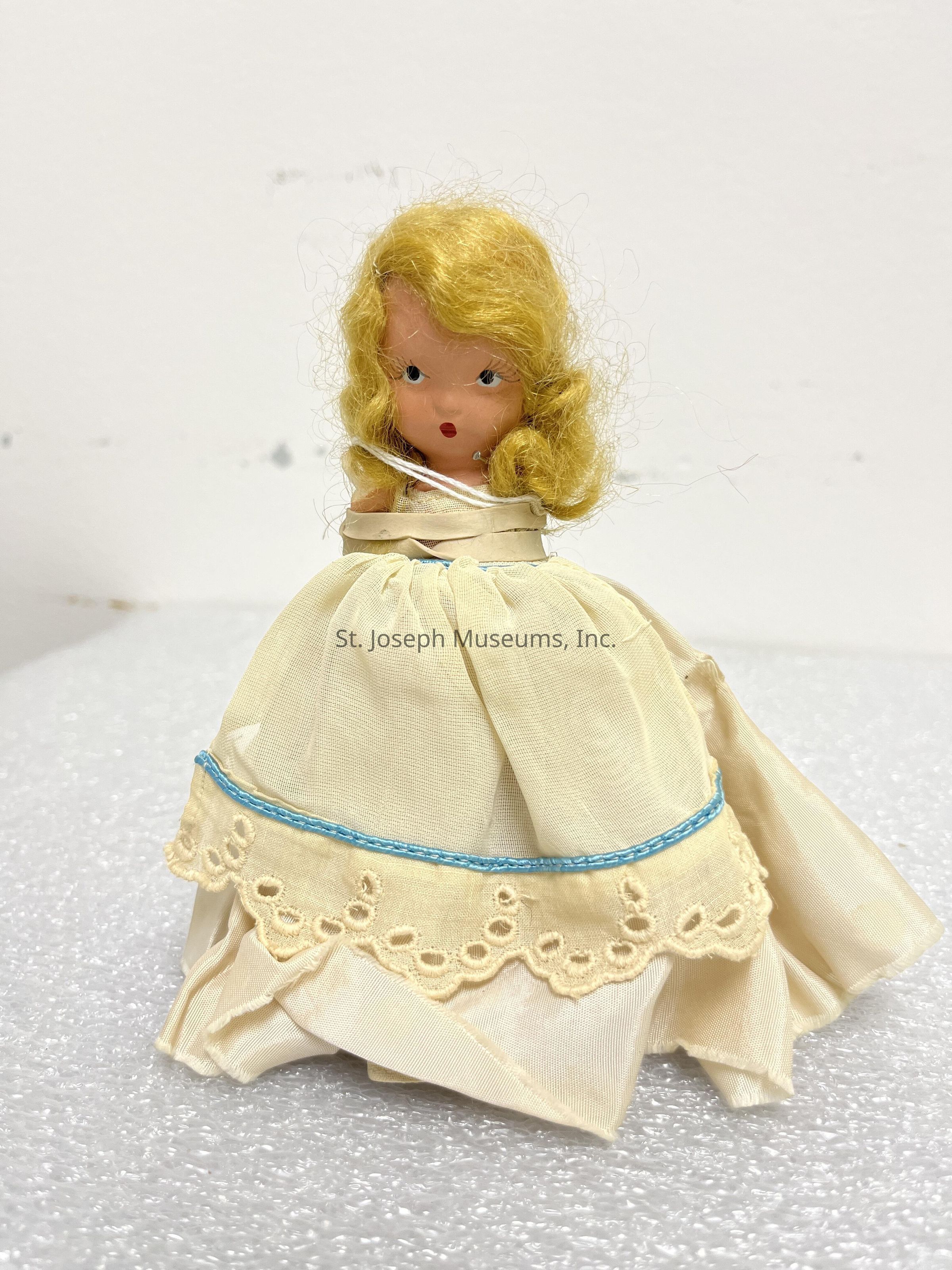 The image depicts a vintage doll with curly blonde hair and a painted face, featuring small, expressive eyes and a subtle red mouth. The doll is dressed in a cream-colored gown with lace detailing near the hem and a blue trim that adds a delicate touch to its attire. The fabric of the dress has a gentle sheen, creating a sense of elegance. The doll appears to be standing on a textured white surface against a plain background, with the simplicity of the setting highlighting its intricate design. The overall aesthetic of the doll conveys a charming, nostalgic quality.