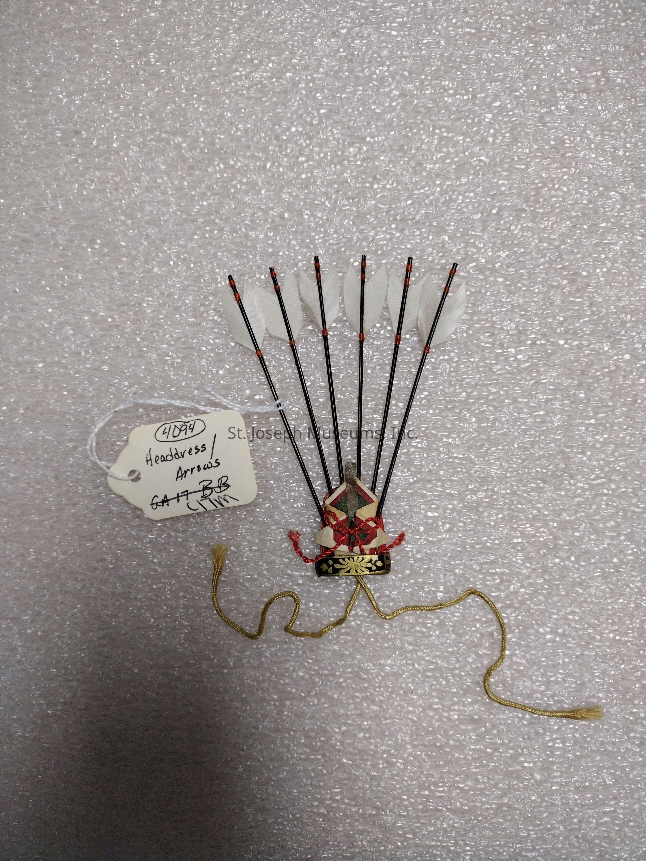 The image shows a traditional ornamental headdress adorned with several vertically arranged arrows that have white feathers at their ends, secured to a decorative base. This base features intricate designs with red and gold accents and is embellished with a red cord tied into a decorative knot. Below the headdress, a gold cord with tassels rests on what appears to be a textured surface, possibly for display or storage purposes. A white tag with handwritten notes identifying the item, possibly for cataloging or exhibition purposes, is attached to the headdress with a string, indicating it as "Headdress/Arrows" with some alphanumeric codes.