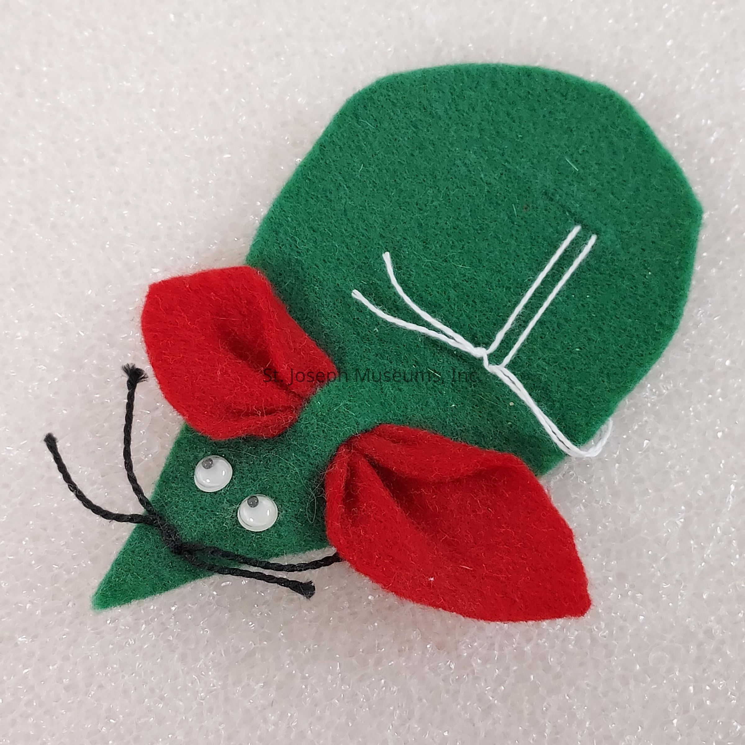 The image features an adorable craft resembling a mouse, made from green and red felt fabric. The body of the mouse is predominantly green and designed in a simplified teardrop shape. Prominent red felt ears are attached, lending contrast and character to the creation. Two small, round, white eyes enhance the playfulness of the mouse’s face. The whiskers are creatively crafted from short, black string, adding to the whimsical aspect of the design. White threads are stitched near the tail region, completing the mouse’s appearance. The craft is laid on a textured, pale surface, highlighting its vibrant colors and simple charm.
