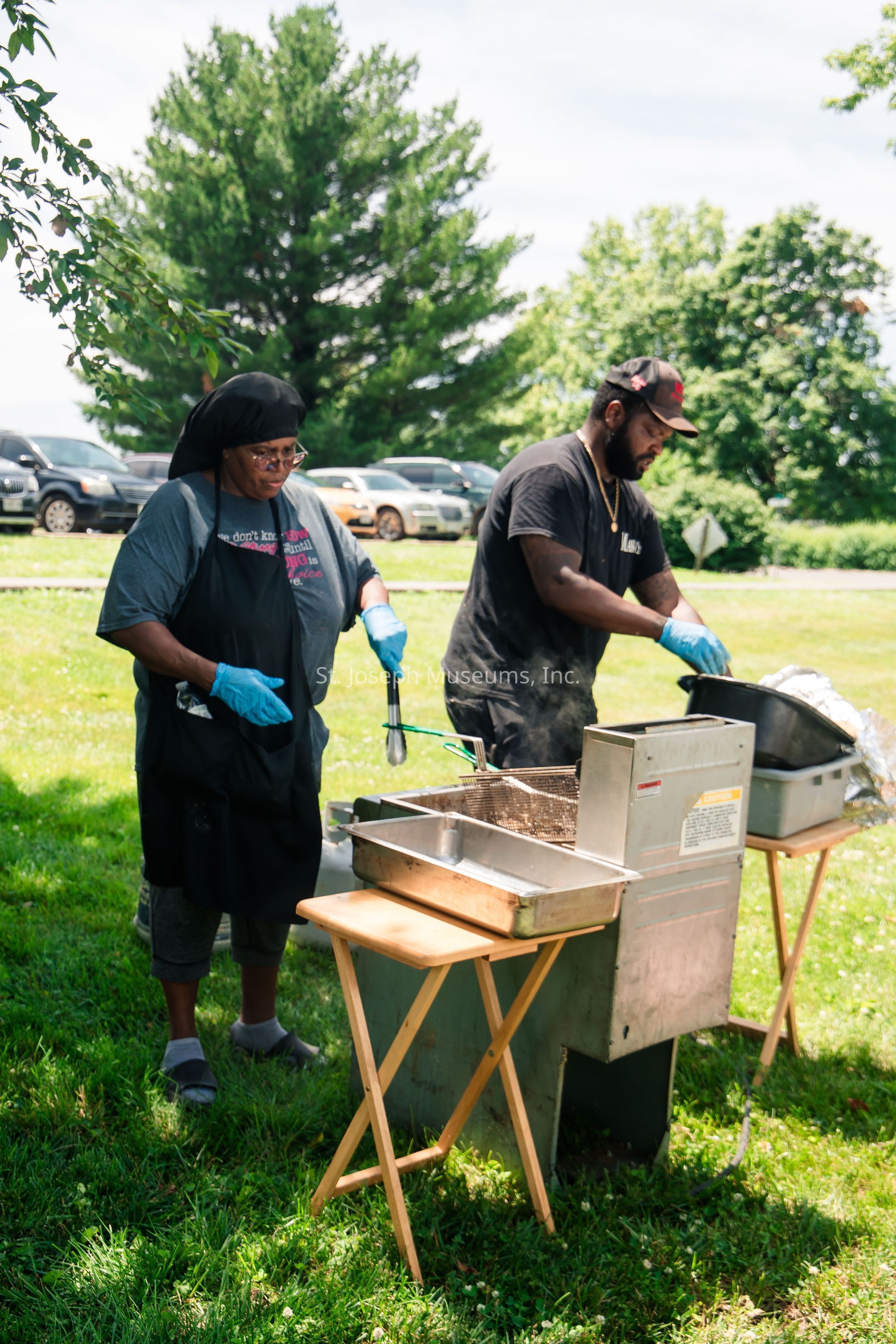 In a serene outdoor setting, two individuals engaged in cooking activities are standing on a lush green lawn, with a variety of cars parked in the background and tall trees providing shade under a bright, partly cloudy sky. Both are wearing protective blue gloves while tending to food in deep metal fryers set on wooden folding tables. The person on the left is wearing a black apron and headscarf, observing the frying process closely, holding a pair of metal tongs. Beside them, another individual with a black cap and T-shirt is also focused on preparing the meal, suggesting a shared effort in a community event or picnic, highlighted by the peaceful surroundings and vibrant green grass.