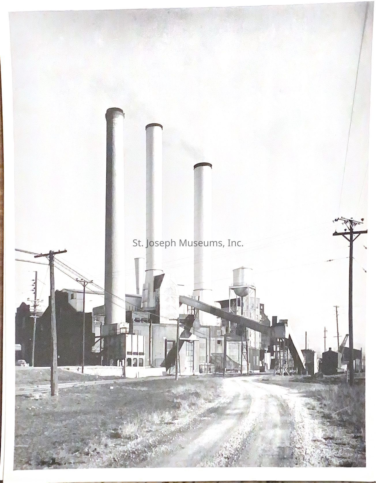 Black-and-white photograph of three tall smokestacks of the St. Joseph Light and Power Company at the intersection of Felix and Main Streets, surrounded by industrial structures, including a conveyor belt system and a water tower, creating a skyline dominated by industrial architecture and machinery.