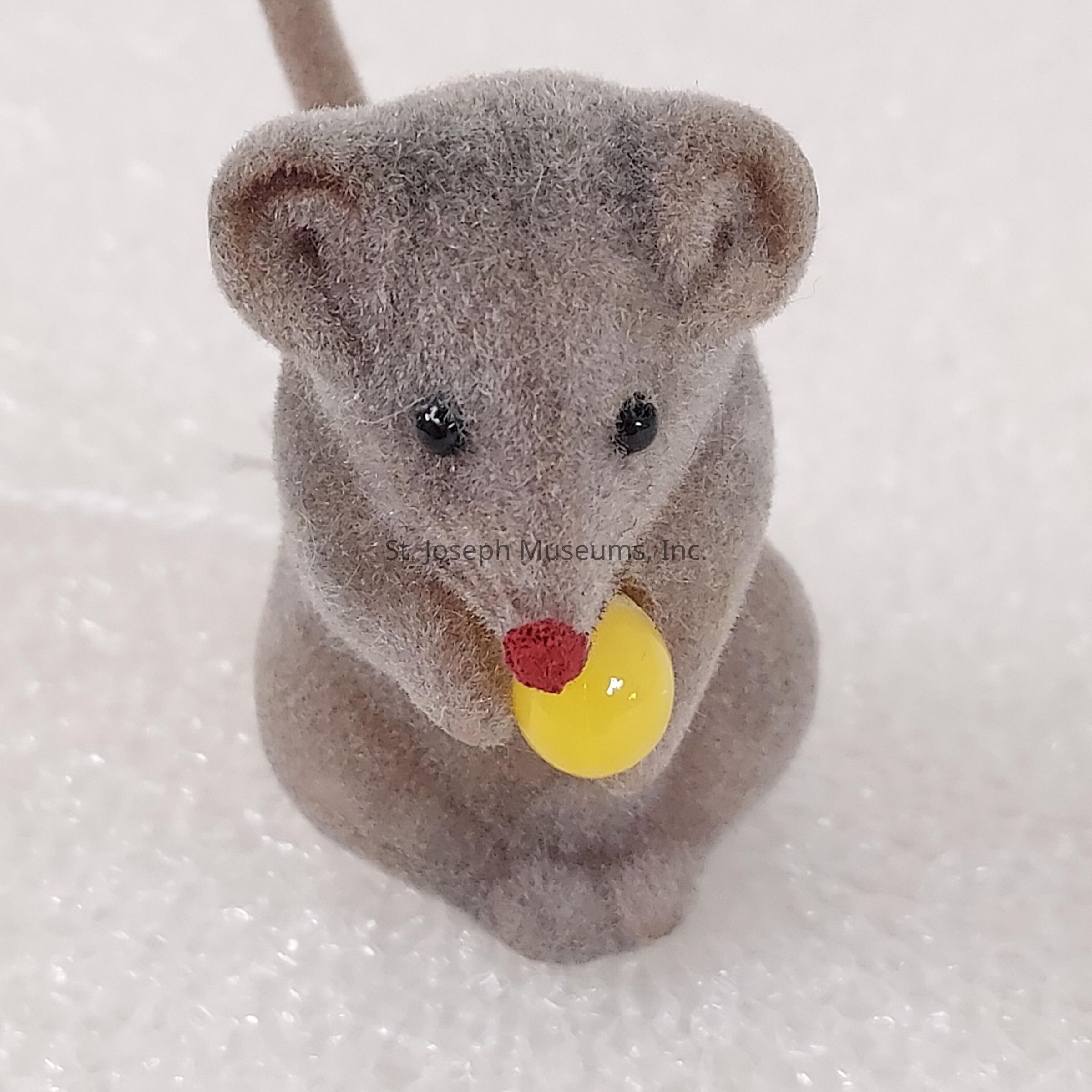 The image shows a small, plush mouse figurine with a soft, fuzzy texture, depicted in a sitting posture. It has a pair of shiny black eyes and a red nose, giving it an endearing and lifelike appearance. The mouse is holding a bright yellow object, possibly representing a piece of cheese or a fruit, which contrasts vividly against its gray body. The surface the mouse is sitting on appears white and textured, possibly resembling snow or a fabric material. This combination of elements creates a charming and whimsical scene with a focus on the mouse's expressive features and colorful accessory.