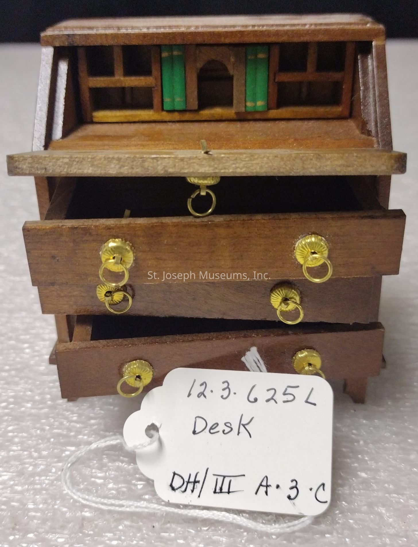 The image showcases a wooden miniature desk with intricate detailing, featuring several small drawers adorned with golden ring handles. The top part of the desk has compartments, some of which contain miniature green books. A handwritten tag attached to the desk reads "12.3.625L Desk DH/III A.3.C." The desk is placed on a textured surface, and its craftsmanship suggests it may be used as a model or for decorative purposes. The drawers appear slightly open, hinting at functional space within the miniature design.