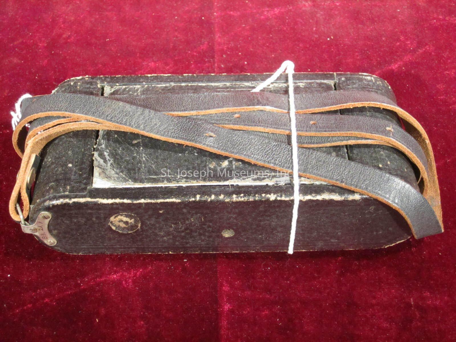 The image shows an old, worn black case with visible signs of aging, such as scuff marks and faded edges, resting on a rich red textured fabric background; the case appears to be rectangular with a rough surface, and it is held together by a leather strap that is looped around it several times, along with a piece of white string tied across the middle, giving it a sense of rustic charm and history, suggesting it could have been used for storing or transporting items, possibly of vintage or antique value, evoking a sense of curiosity about its past and what it may have contained or the journeys it has been on throughout the years.