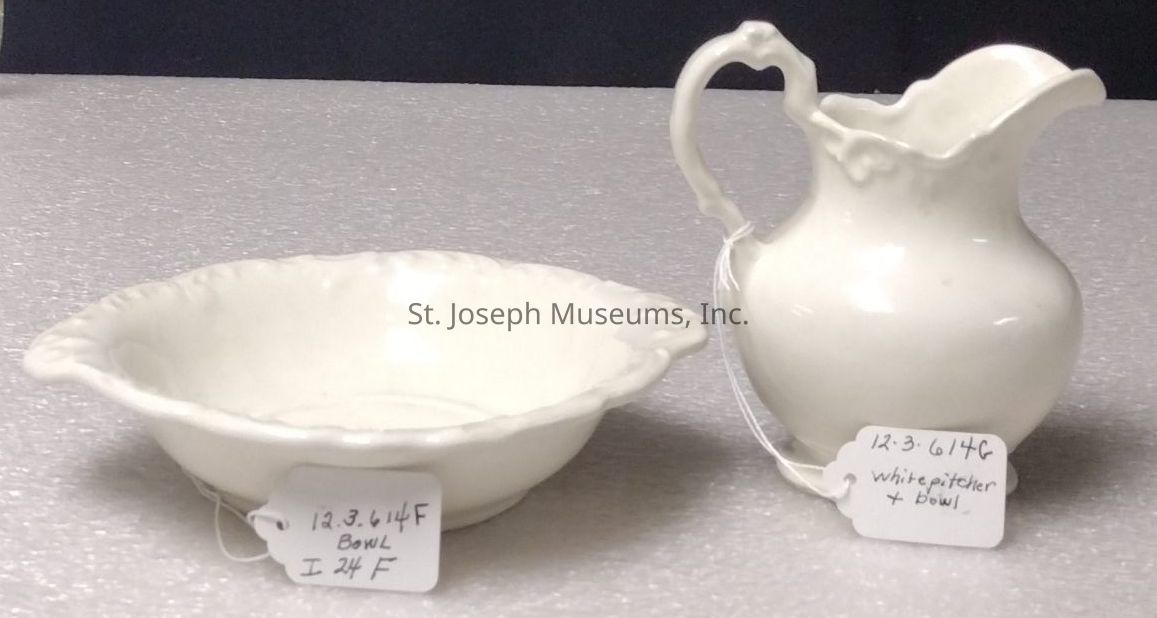 The image shows two pieces of white ceramic tableware set against a textured surface: a bowl and a pitcher. The bowl, positioned on the left, has an ornate rim and is accompanied by a small tag tied to it, labeled with handwritten text reading "12.3.614F Bowl." On the right side, there is an elegant pitcher with a gracefully curved handle, which also features a handwritten tag reading "12.3.614G white pitcher + bowl." Both items are simple yet elegant in design, capturing a classic and sophisticated aesthetic.