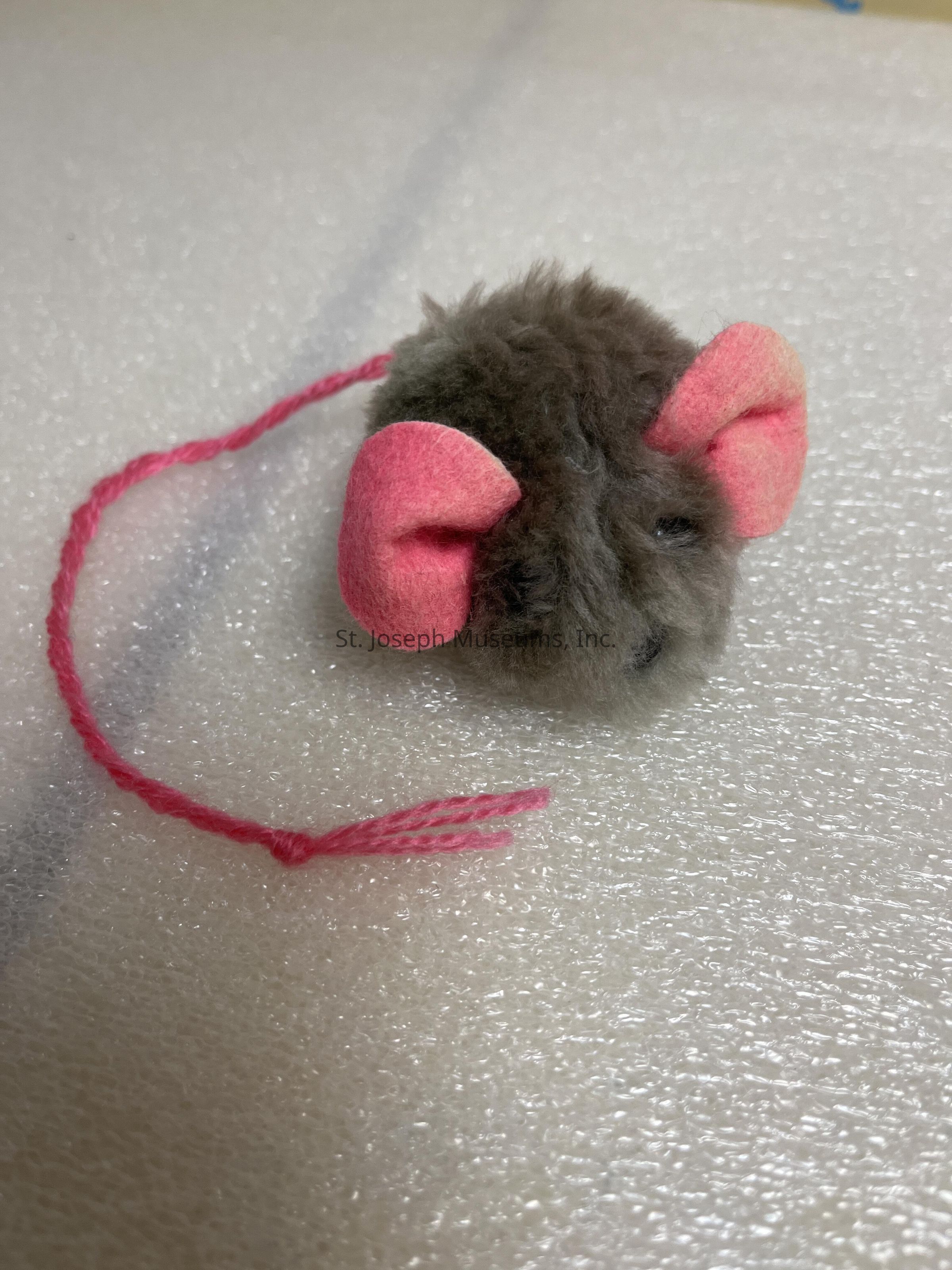 The image shows a small, plush toy mouse with a fluffy, brownish-gray body, featuring two prominent pink ears and a matching slender pink tail, placed on a textured white surface, which likely provides contrast to highlight the toy's soft material and delightful features, suggesting it could be designed for a pet, such as a cat, to play with. The texture of the fur on the toy mouse is evident, giving it a lifelike appearance that might be enticing to animals. The tail made from pink yarn adds a playful touch to the whimsical design, making it eye-catching and fun, aligning with its purpose as a pet toy.