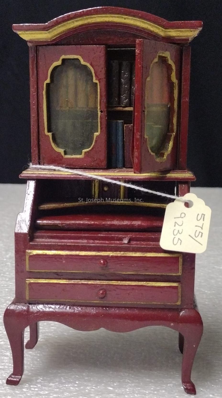 The image showcases a miniature antique-style wooden cabinet painted in a deep red color with golden accents. The cabinet features an arched top and two glass doors, through which a few tiny books can be seen. Below the bookshelf section is a small drop-down desk with a flat surface, adding functionality to the piece. The lower section consists of two drawers with small, rounded knobs. It stands on elegant, curved legs, enhancing its vintage aesthetic. A white string is attached to the cabinet, holding a small beige tag marked with the numbers "575/9235," indicating a form of identification or cataloguing. The background is a subtle, neutral color, ensuring that the intricate details of the cabinet are prominently highlighted.