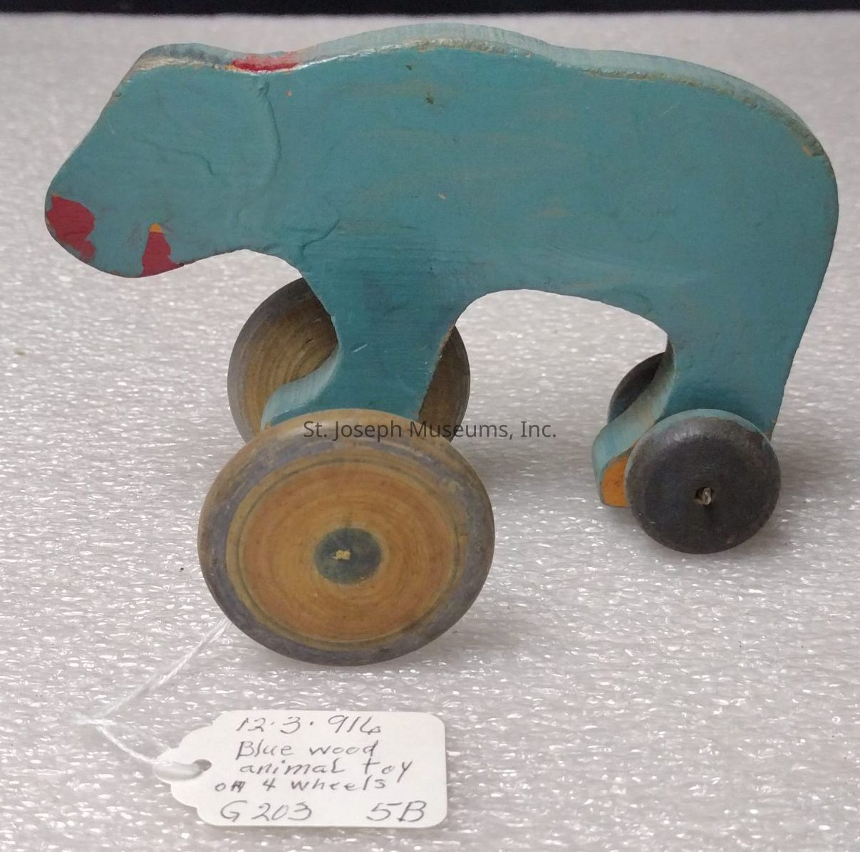 The image depicts a vintage wooden toy in the shape of an animal, likely a bear, painted in blue with a hint of red around its mouth and snout. The toy is designed on four wheels, allowing it to be pushed or rolled around. The wheels are cylindrical and resemble natural wood with concentric circles visible, signifying that they might have been manually crafted. A small paper tag is attached to the toy with a string, inscribed with handwritten text detailing "12.3.914 Blue wood animal toy on 4 wheels G203 5B." The background is a textured, light-colored surface, possibly some sort of padding or foam, which complements the vintage aesthetic of the toy. This antique item showcases a simple yet charming design, reminiscent of early handmade toys.